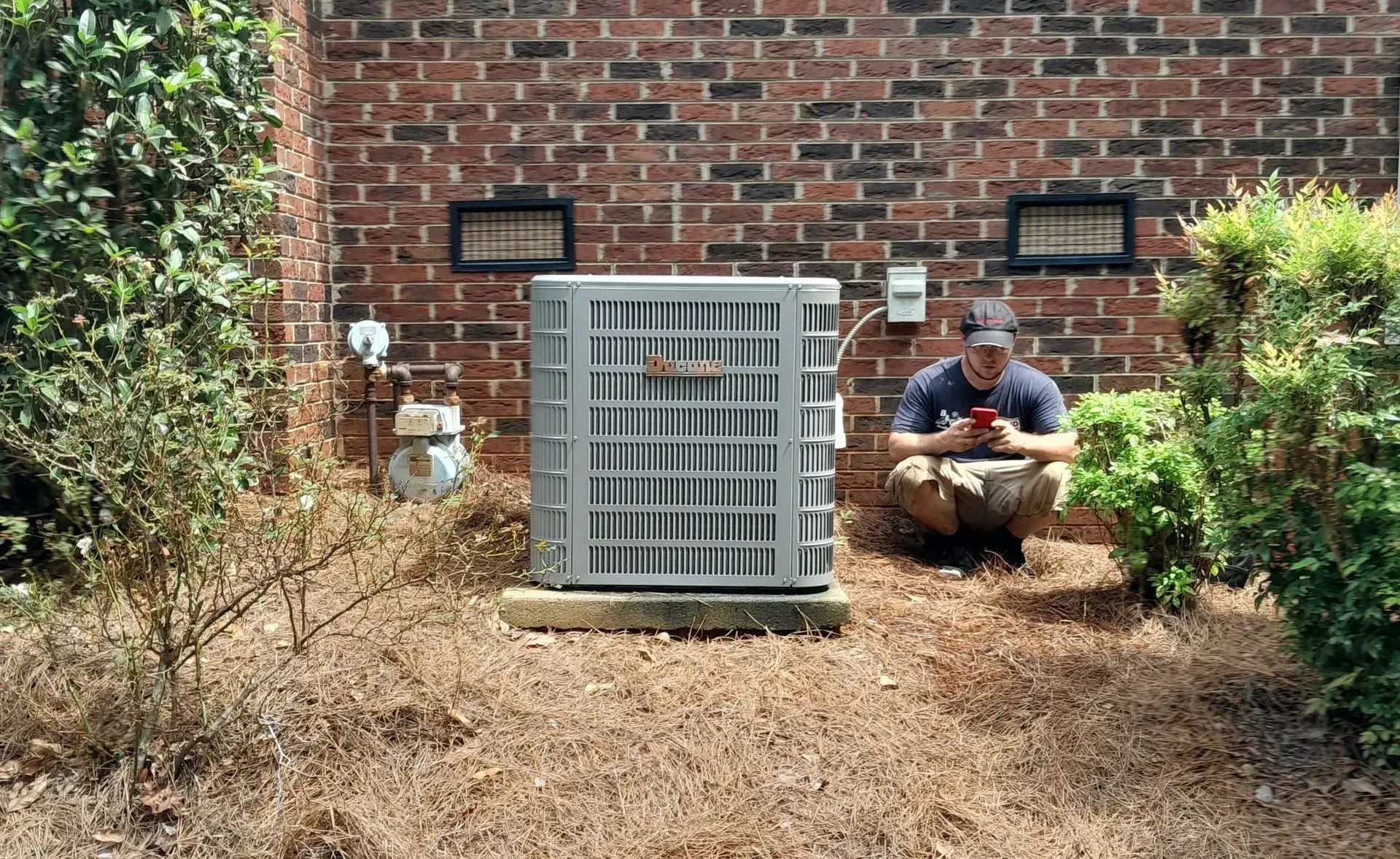 Technician crouches beside outdoor HVAC unit against a brick wall, inspecting equipment.