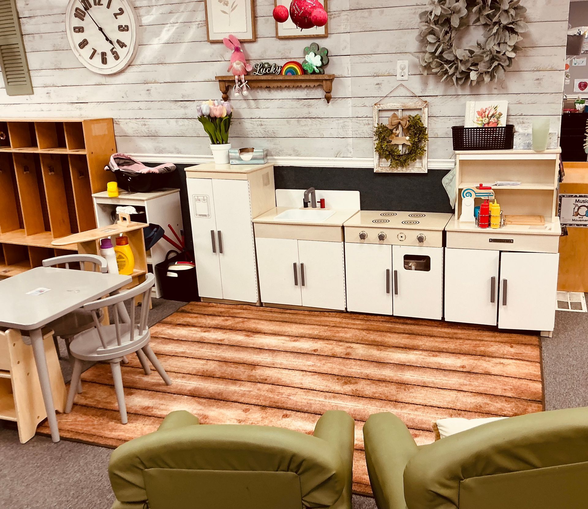 Play kitchen in a classroom with white cabinets and a rug. Table and chairs in front with green chairs.