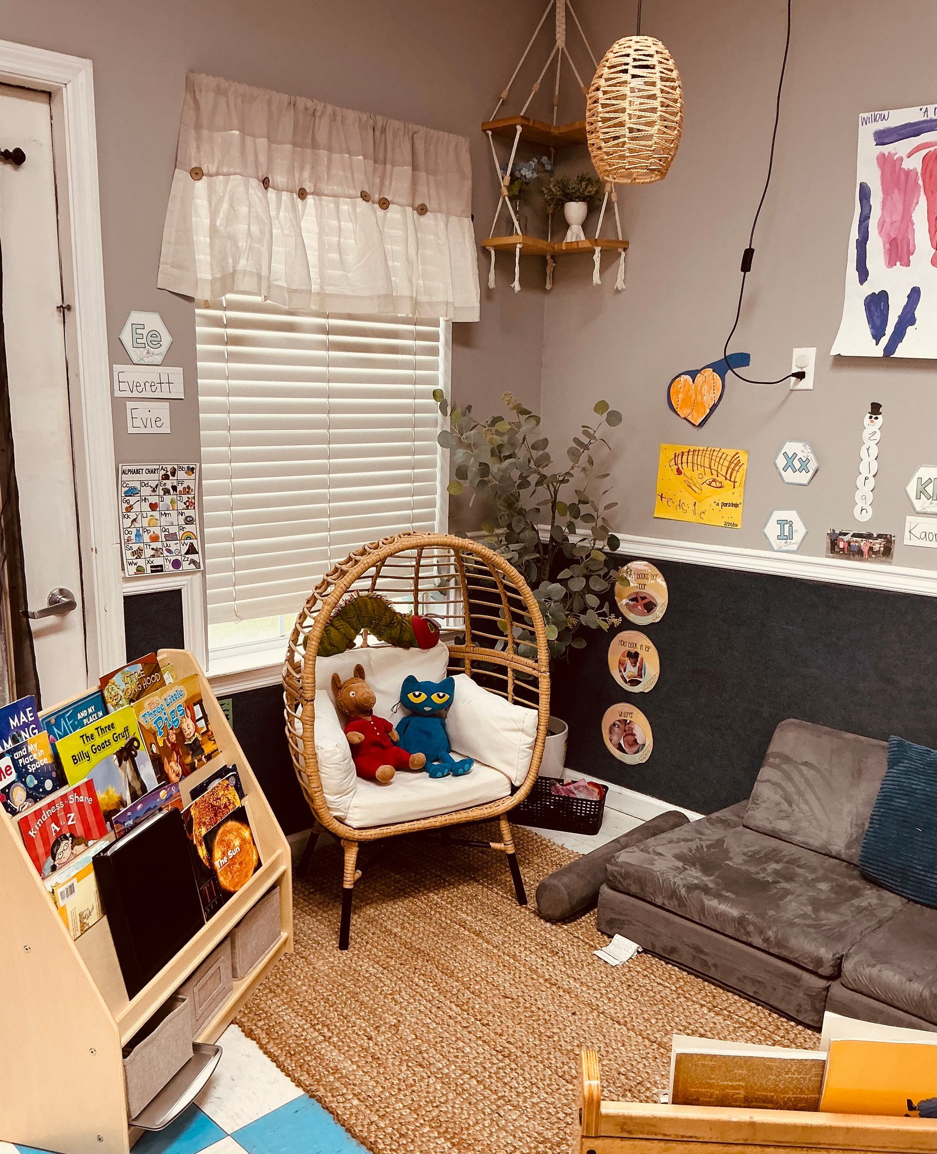 Cozy reading nook with a rattan chair, bookshelf, and sofa. Artwork and hanging decor adorn the gray walls.