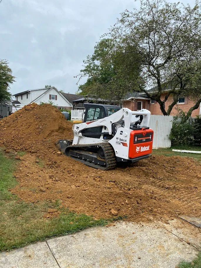 White Bobcat skid steer by a dirt mound in a residential yard, with a tree and houses in the background