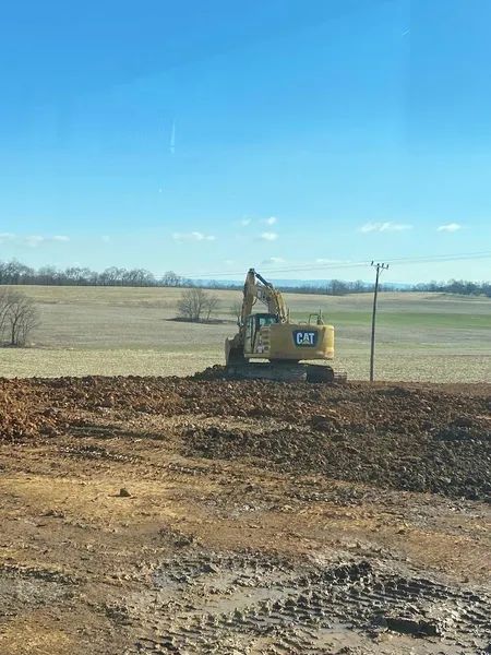 Yellow excavator on a dirt field under a clear blue sky, with open farmland in the background.