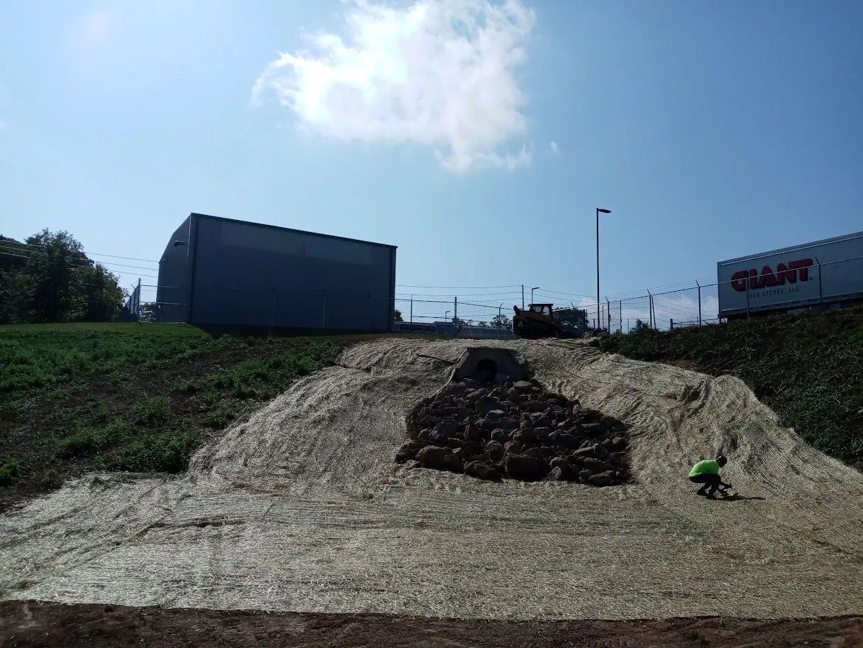 Gravel mound with dark debris on a roadside embankment under a blue sky, with buildings in the background