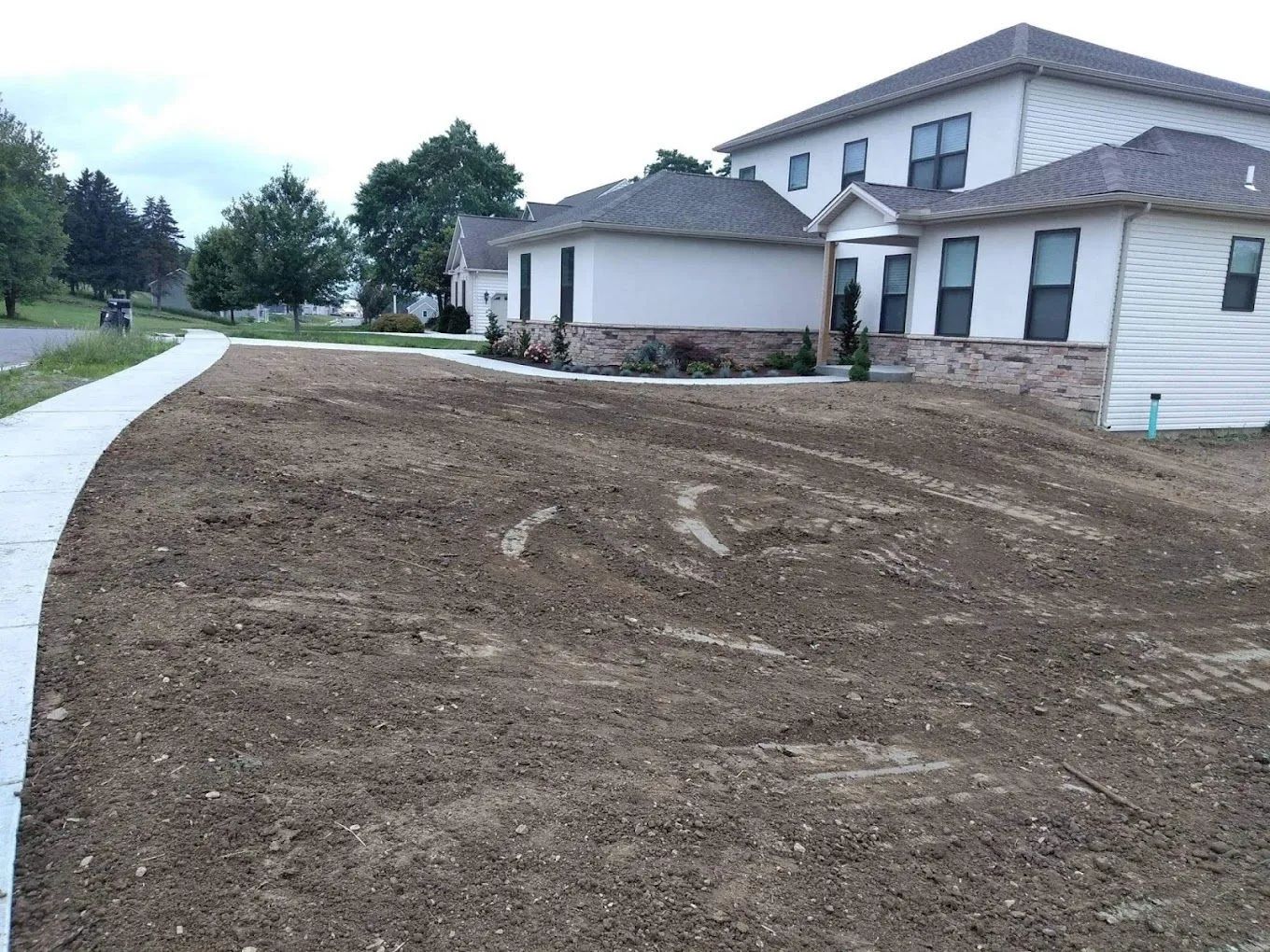 Freshly graded dirt lot beside a curved sidewalk and white house under construction