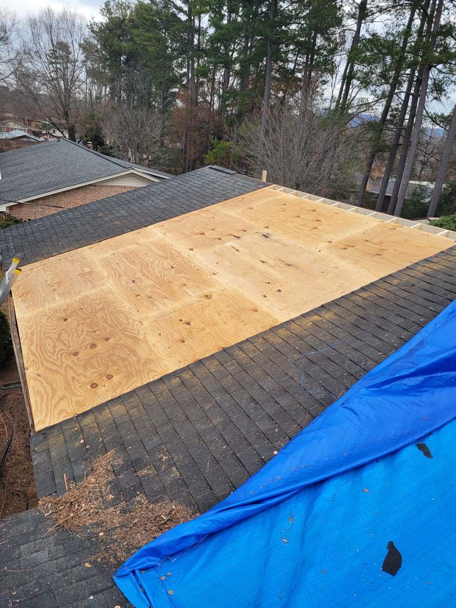 A large piece of plywood is sitting on top of a roof next to a blue tarp.