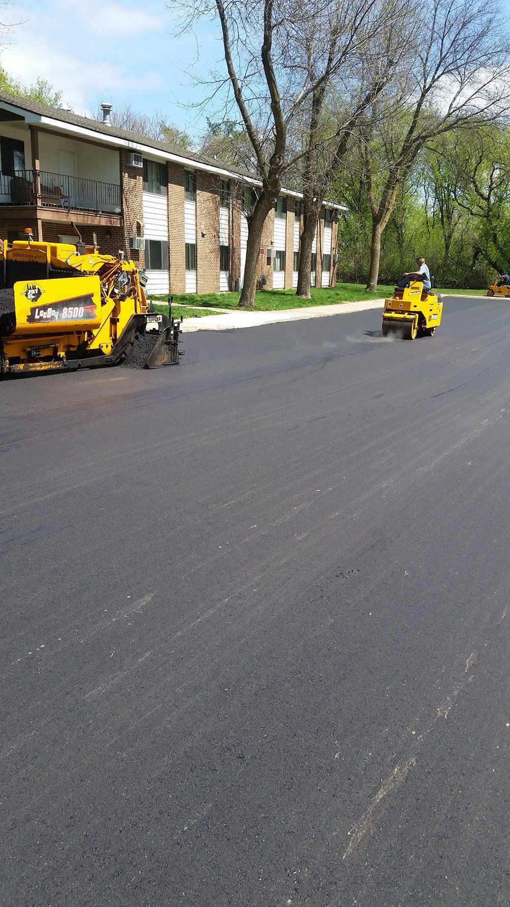 Yellow paving machinery works on fresh black asphalt in front of a multi-story apartment building on a sunny day.