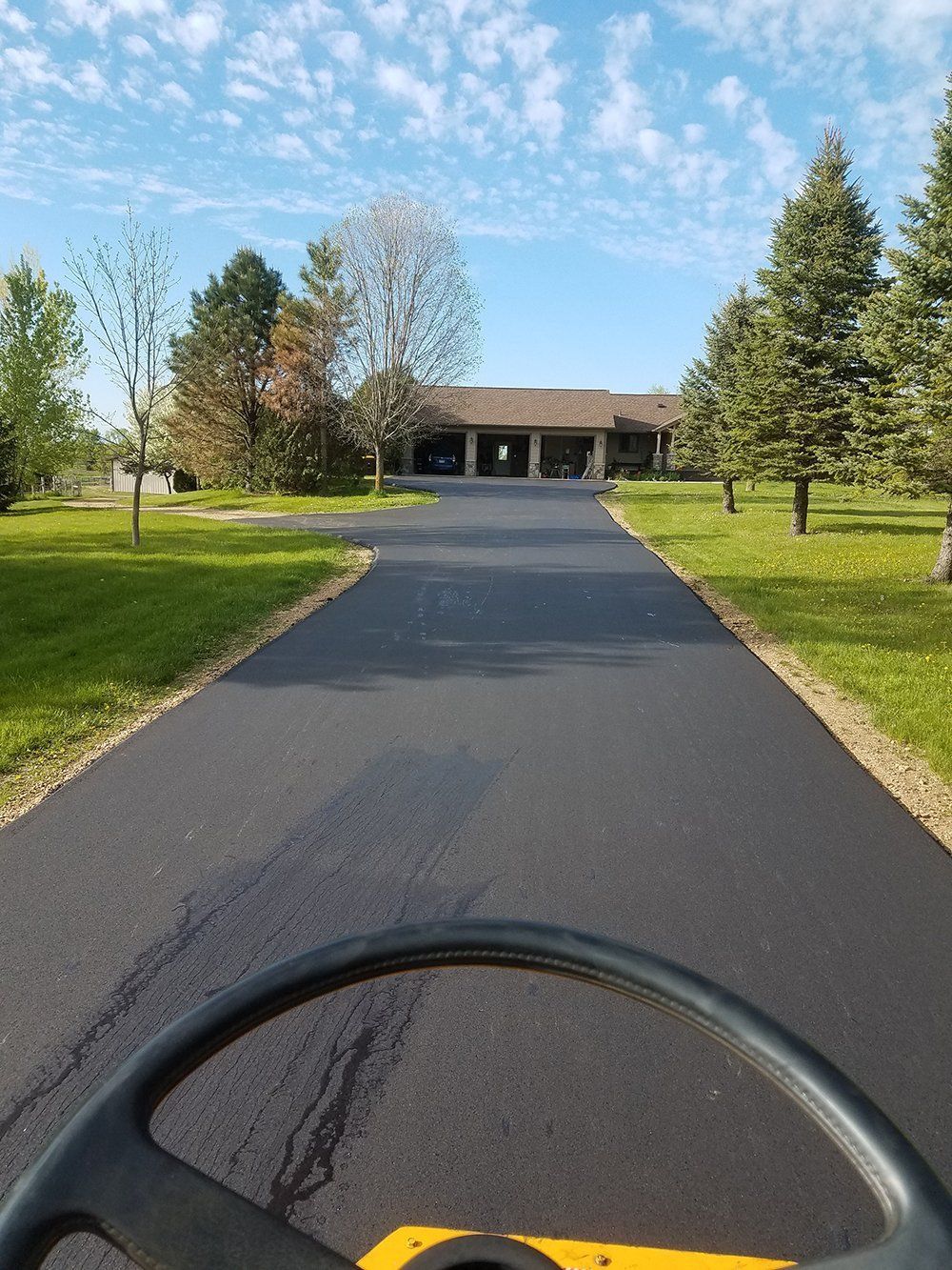 A view from a construction vehicle looking down a freshly paved asphalt driveway toward a house under a sunny blue sky.