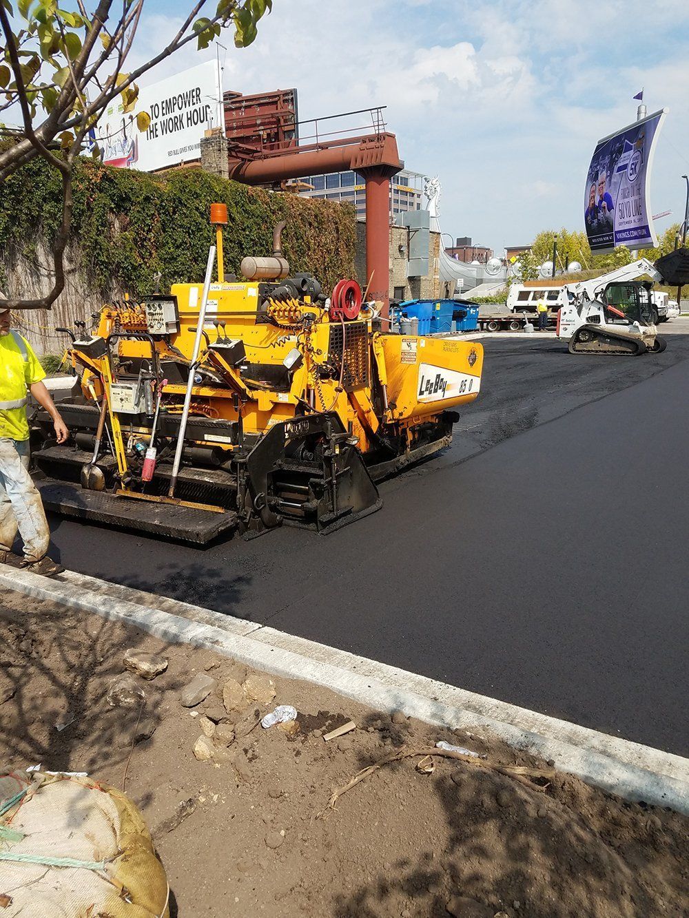 A yellow asphalt paving machine works on a newly laid dark road surface under a clear sky.