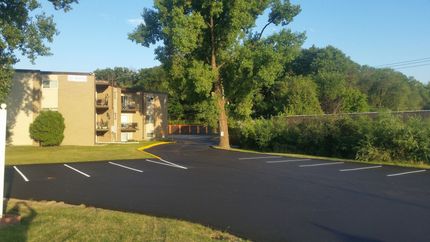 Newly paved parking lot in front of a tan apartment building, surrounded by green trees and grass on a sunny day.
