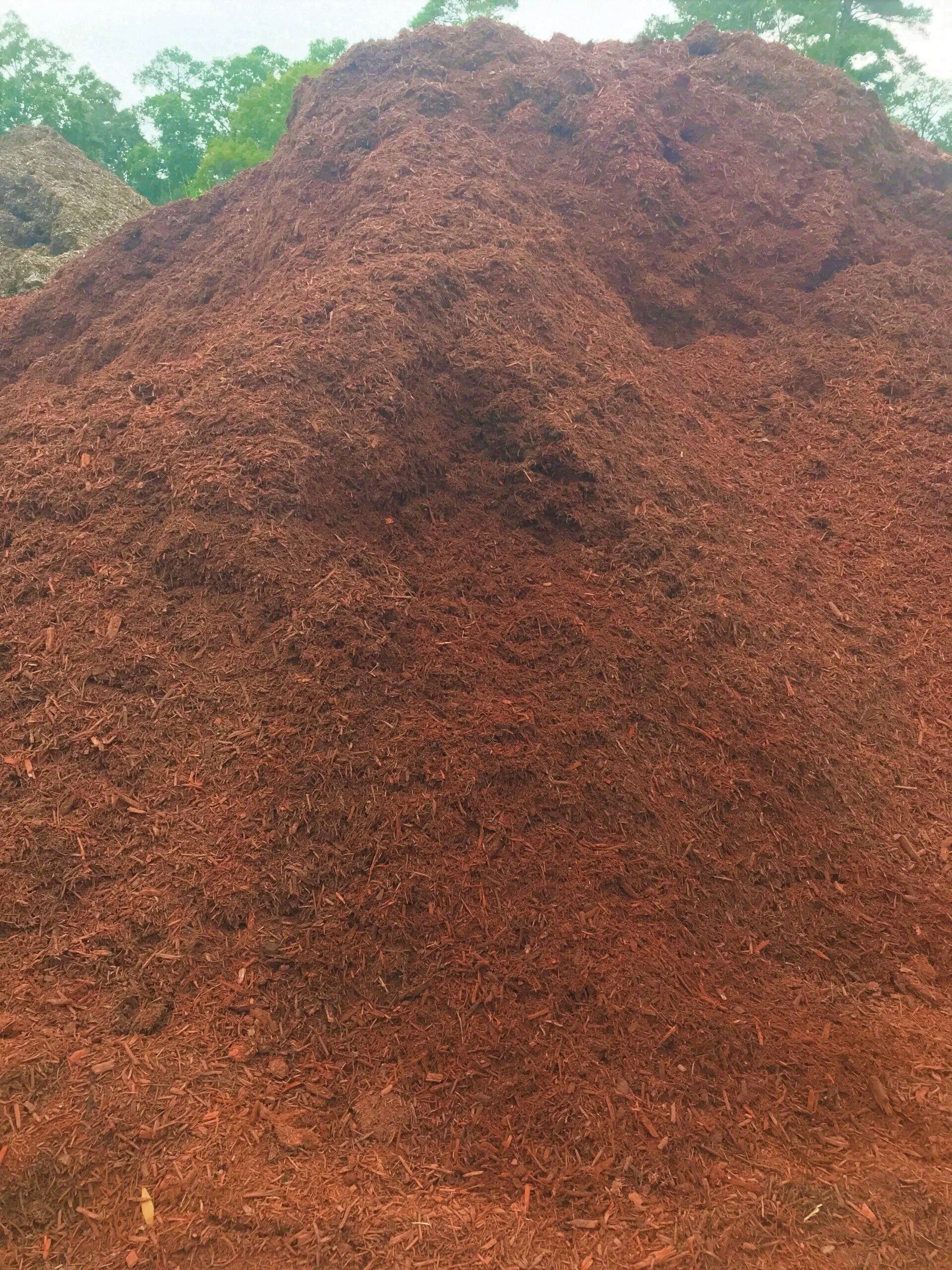Large reddish-brown mound of wood chips or mulch outdoors, with green trees in the background