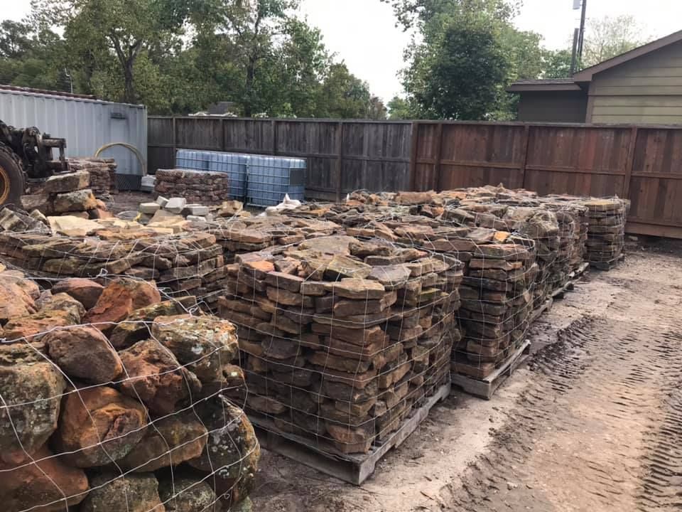 Stacks of weathered bricks and stone pallets in a dirt yard beside a wooden fence.