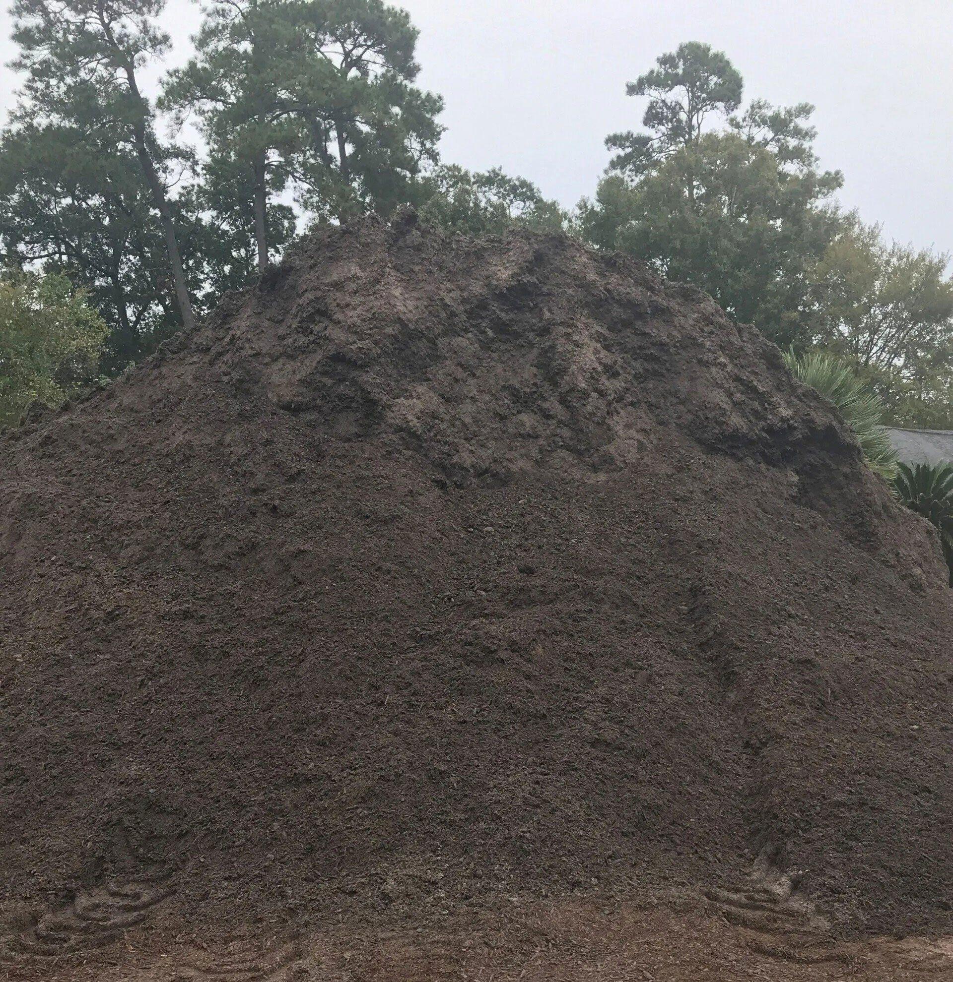 Large dark pile of soil or compost outdoors, with trees in the background.