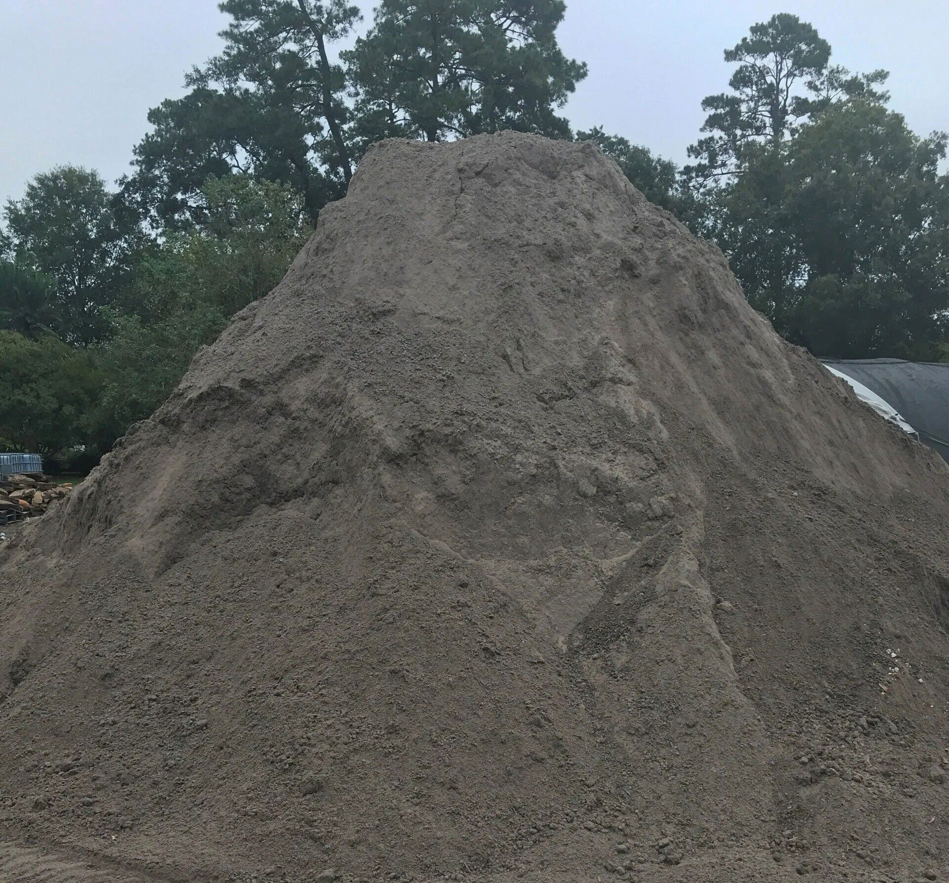 Large mound of gray gravel or sand outdoors with trees in the background