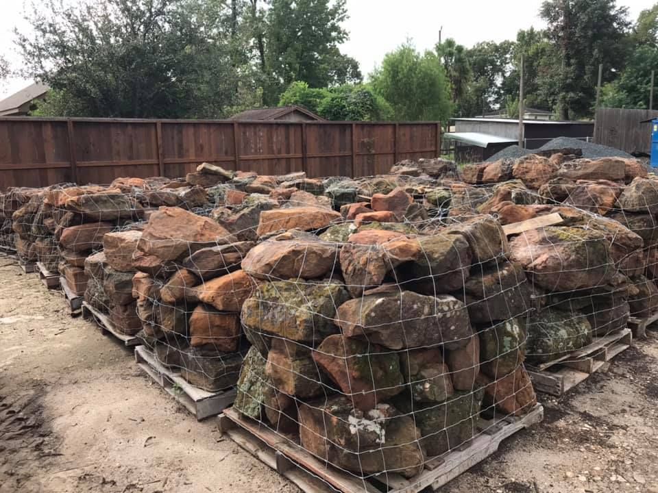 Stacked firewood and logs wrapped in mesh on pallets in a fenced outdoor yard