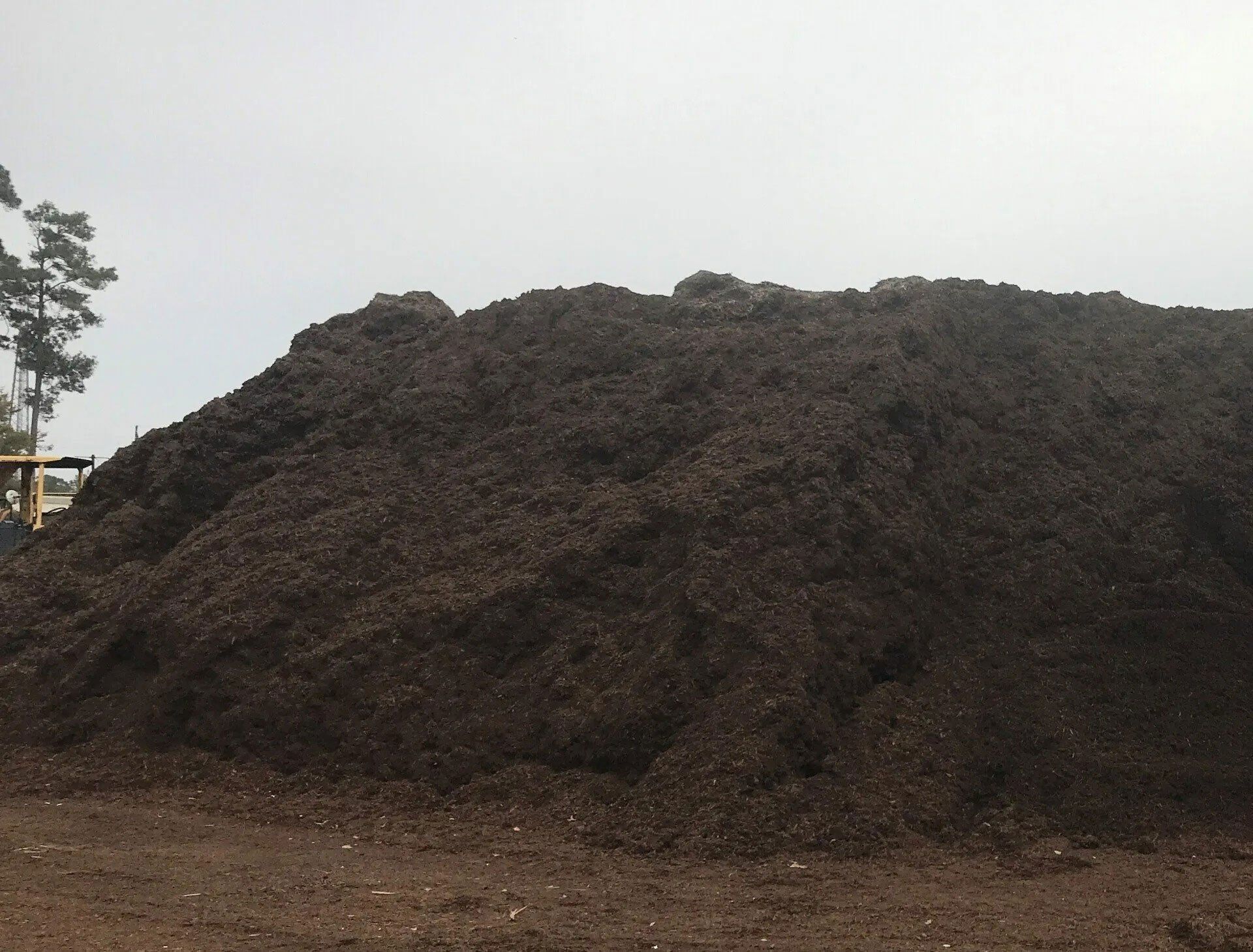 Large pile of dark soil or compost outdoors on bare ground under an overcast sky
