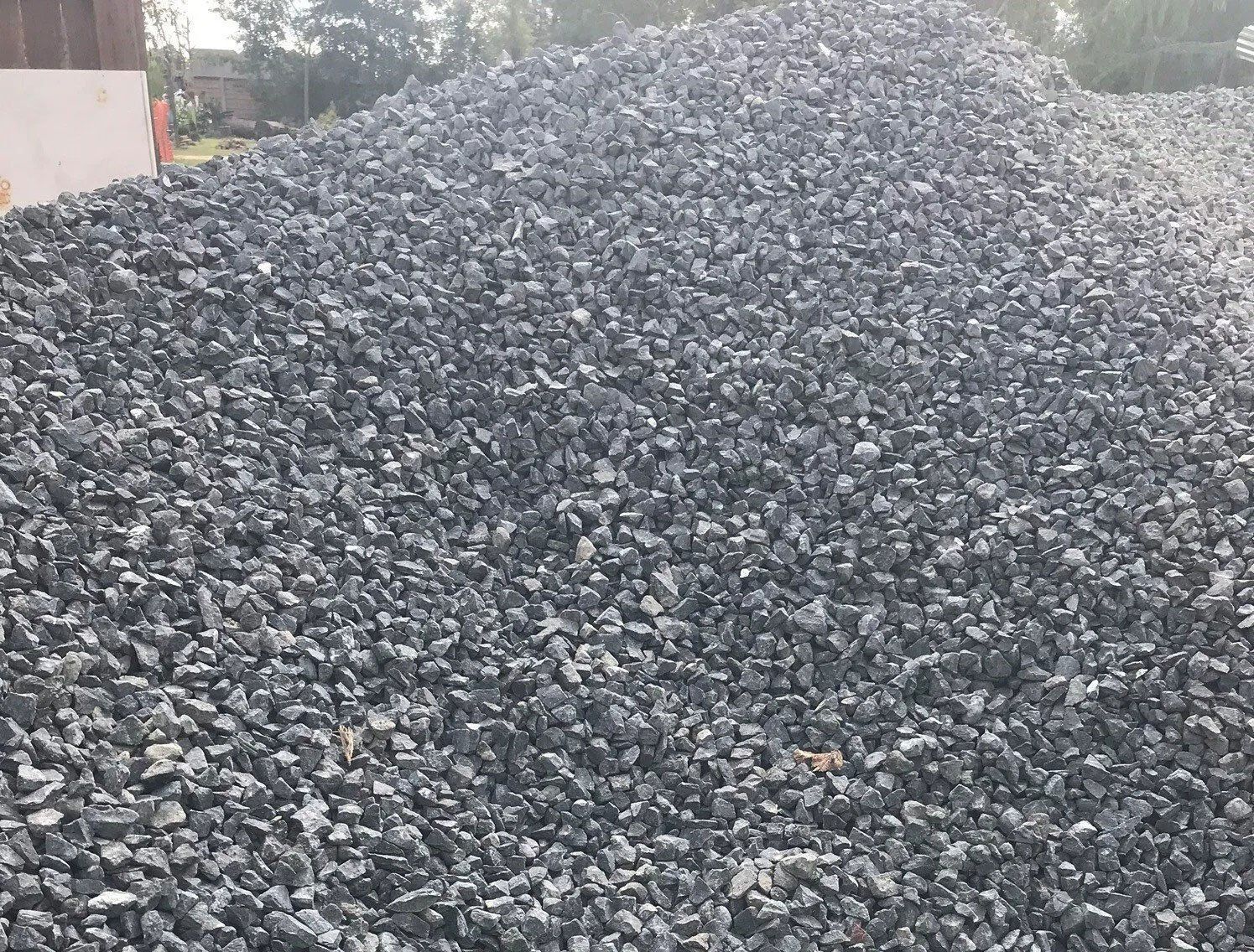 Large pile of gray crushed stones or gravel outdoors with trees in the background