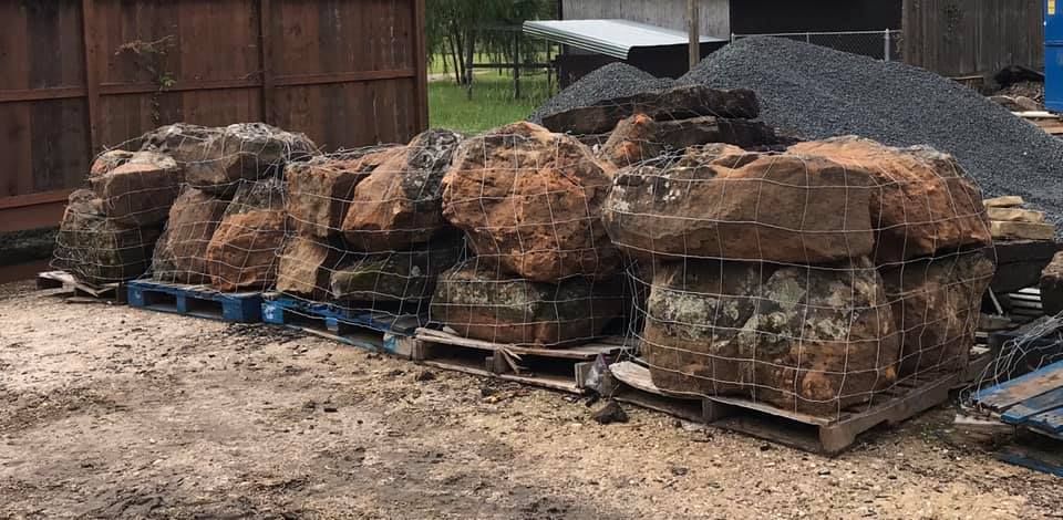 Large pile of rocks and boulders stacked on blue pallets beside a wooden fence.
