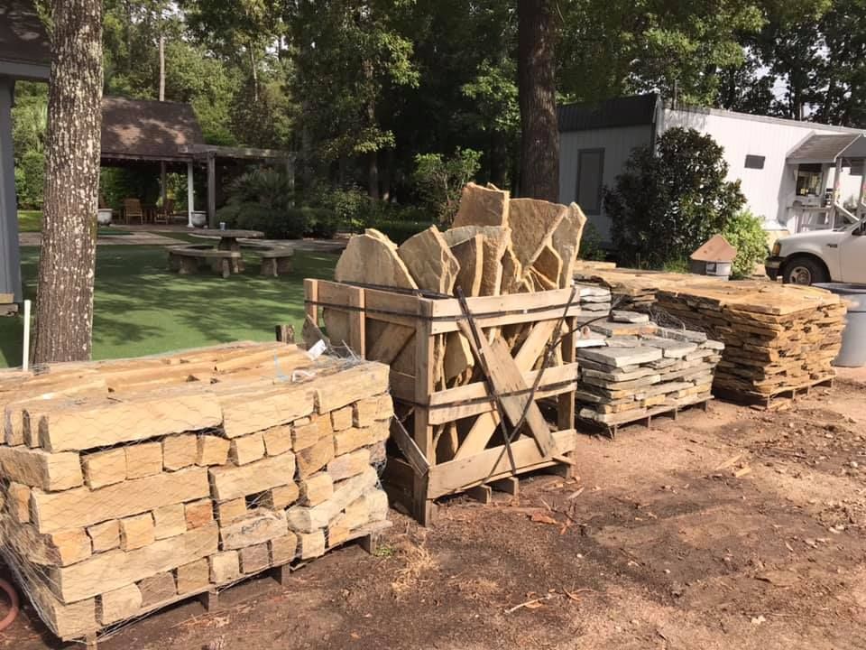 Stacks of firewood and stone slabs in a yard beside a house and trees.