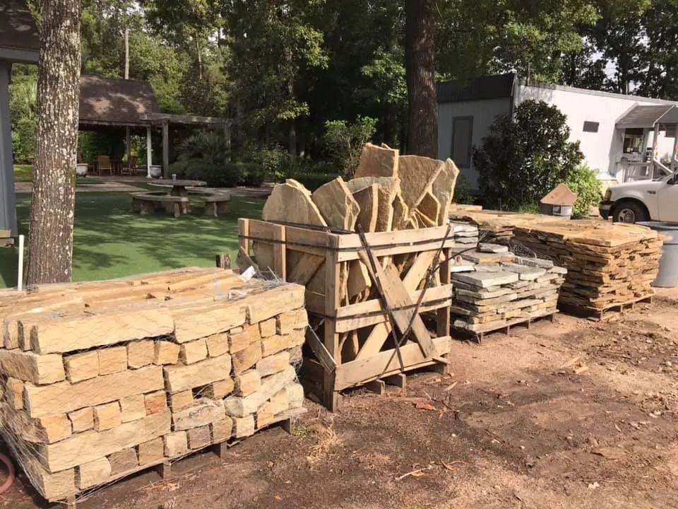 Stacks of cut stone blocks and slabs on pallets in a wooded yard, with a white truck in the background