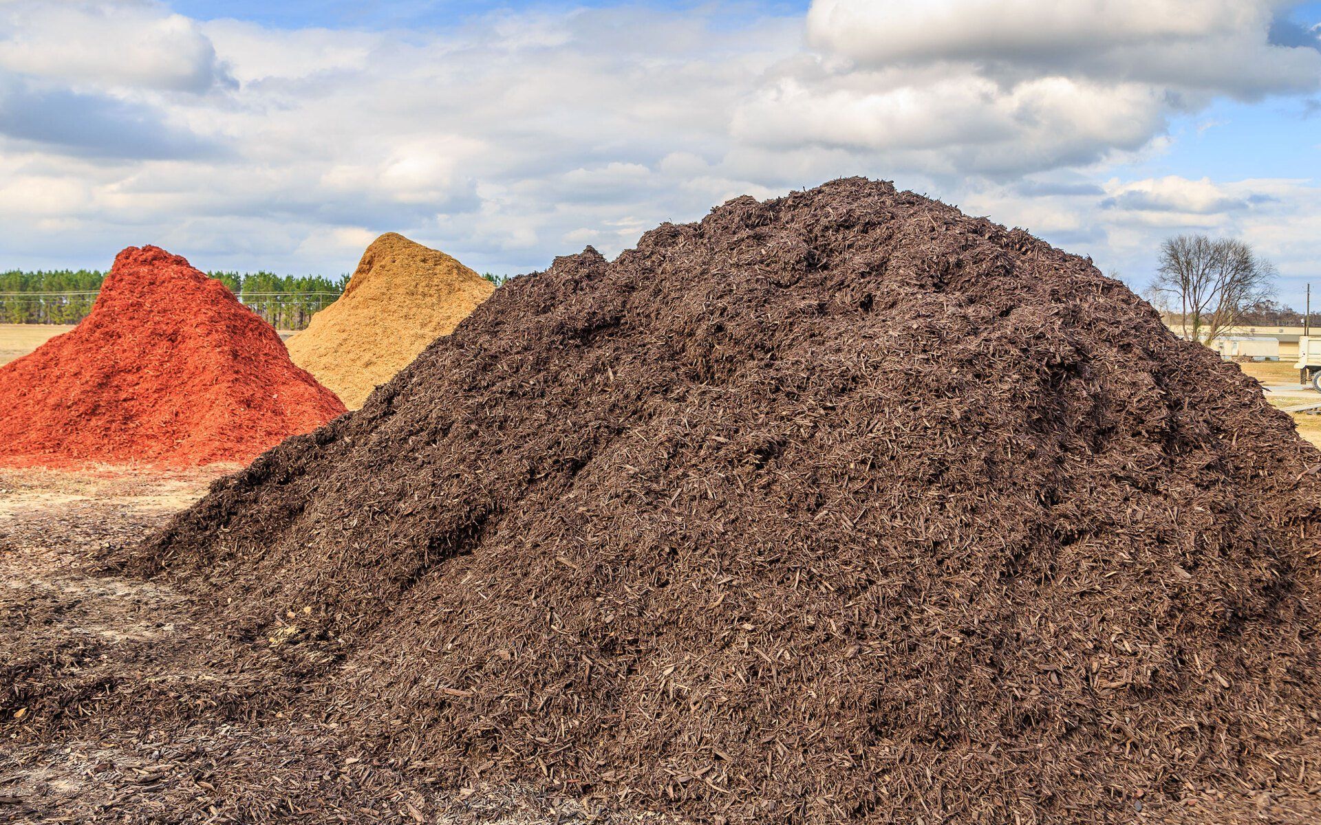 Three mulch piles in red, tan, and dark brown stacked outdoors under a cloudy sky