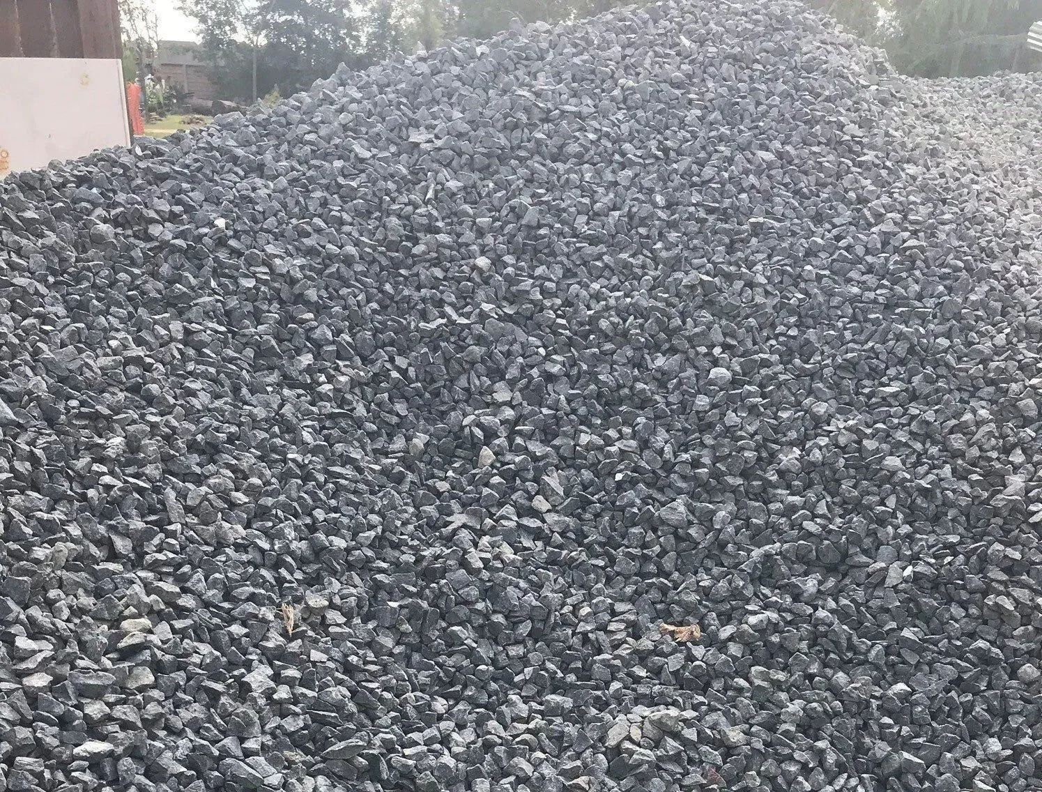 Large pile of gray crushed stone outdoors, with trees in the background