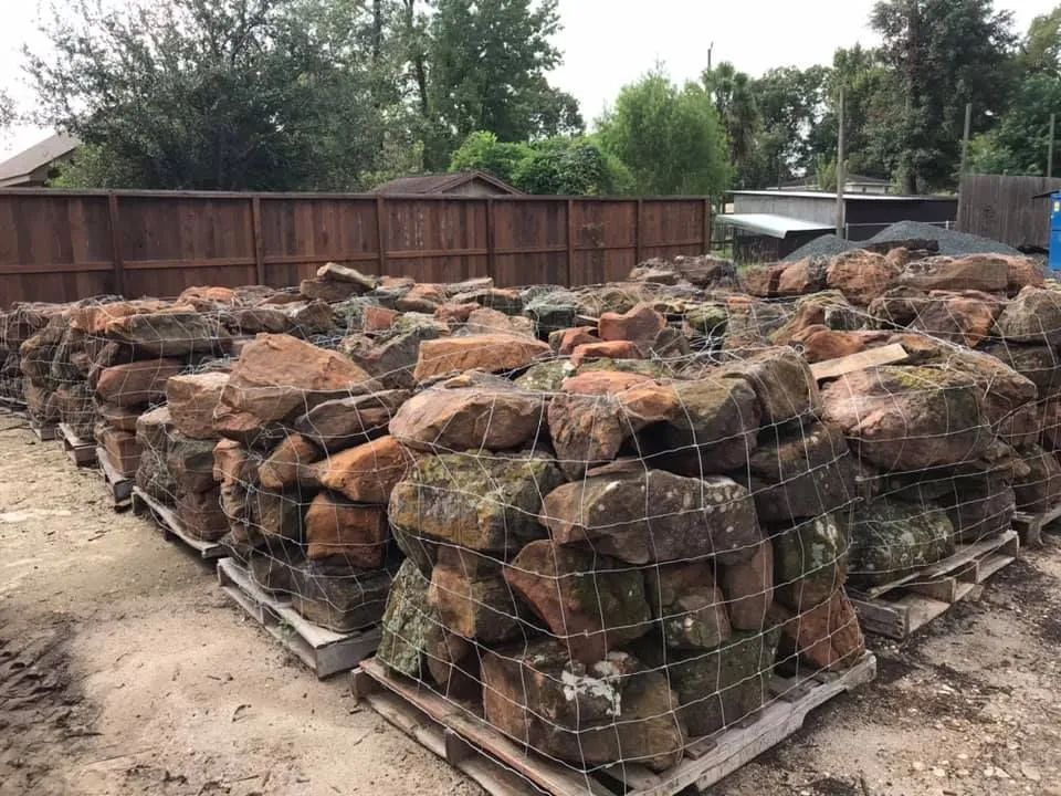 Stacks of firewood bundled on pallets in an outdoor yard, with a wooden fence and trees behind.