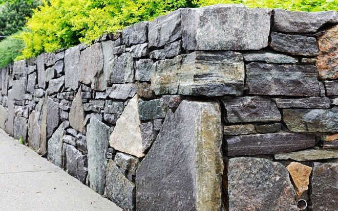 Stone retaining wall beside a sidewalk, with green foliage above.