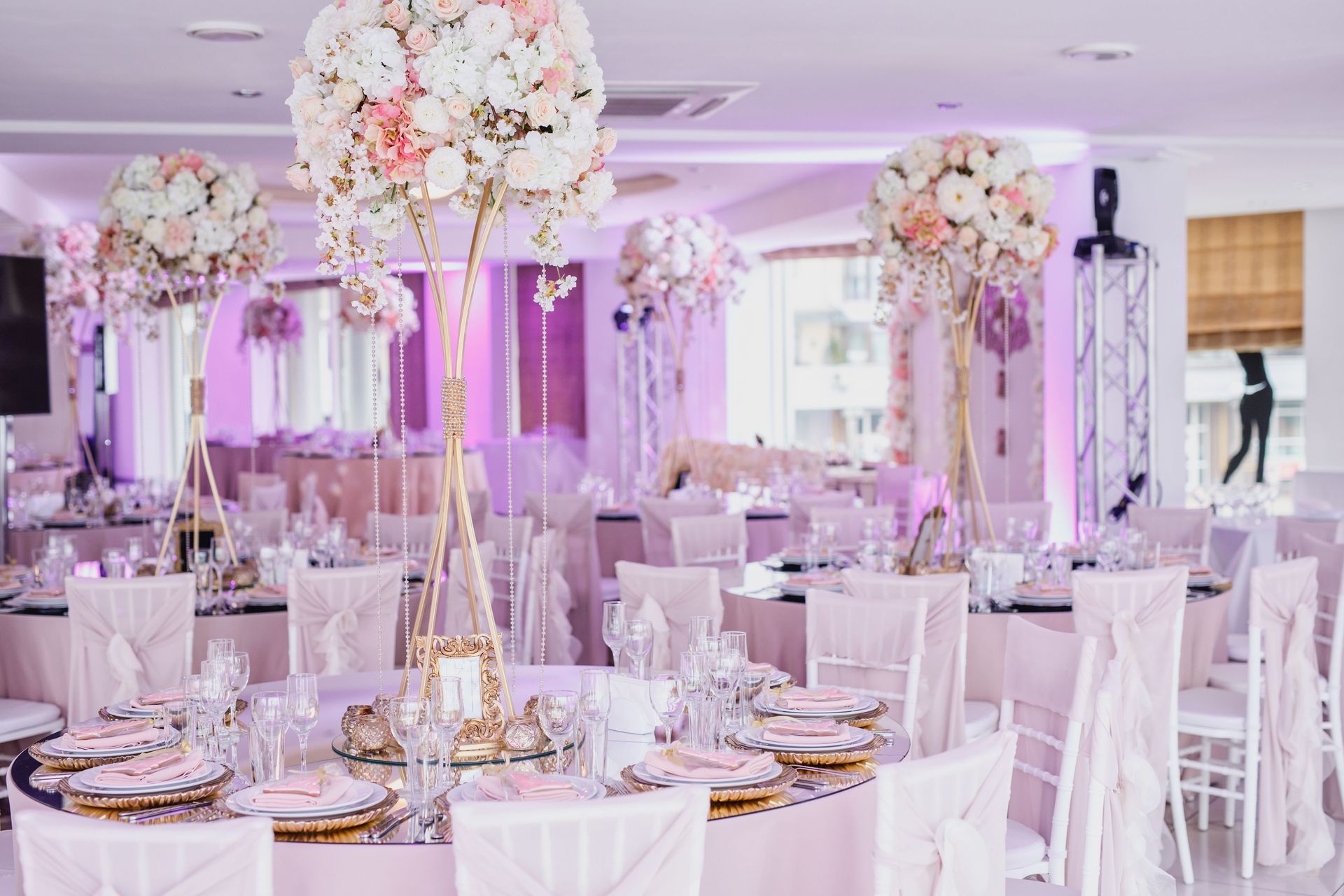 A banquet hall set for a wedding with round tables, light pink linens, and large floral centerpieces on tall stands.
