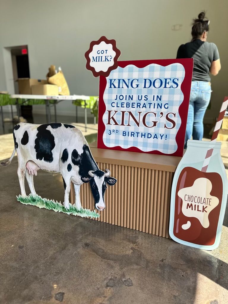 Cow-themed birthday sign reading “King Doss,” with milk bottle cutouts and a cow at a dairy display