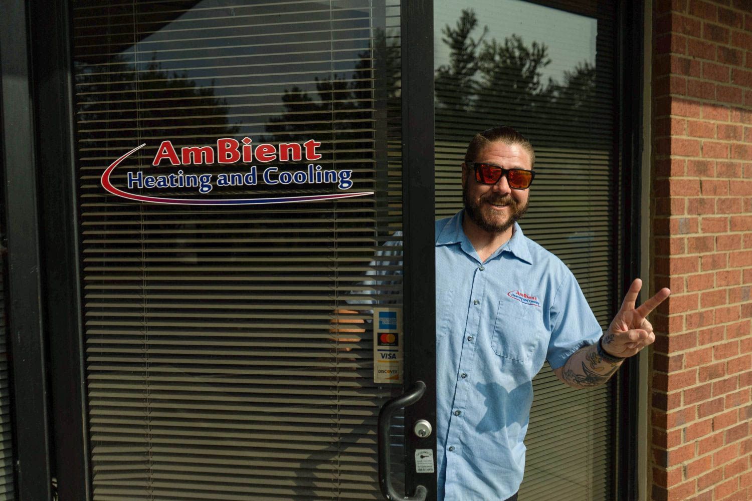 Man standing in a doorway, smiling and making a peace sign outside an office with blinds and a logo sign.