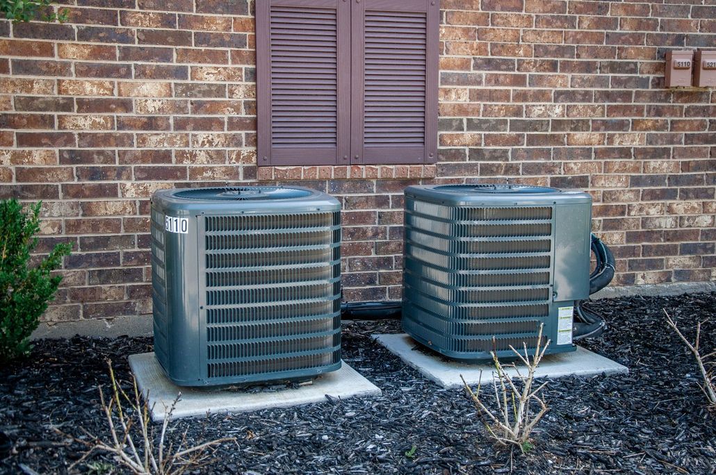 Two outdoor air conditioner units beside a brick house wall and window shutters