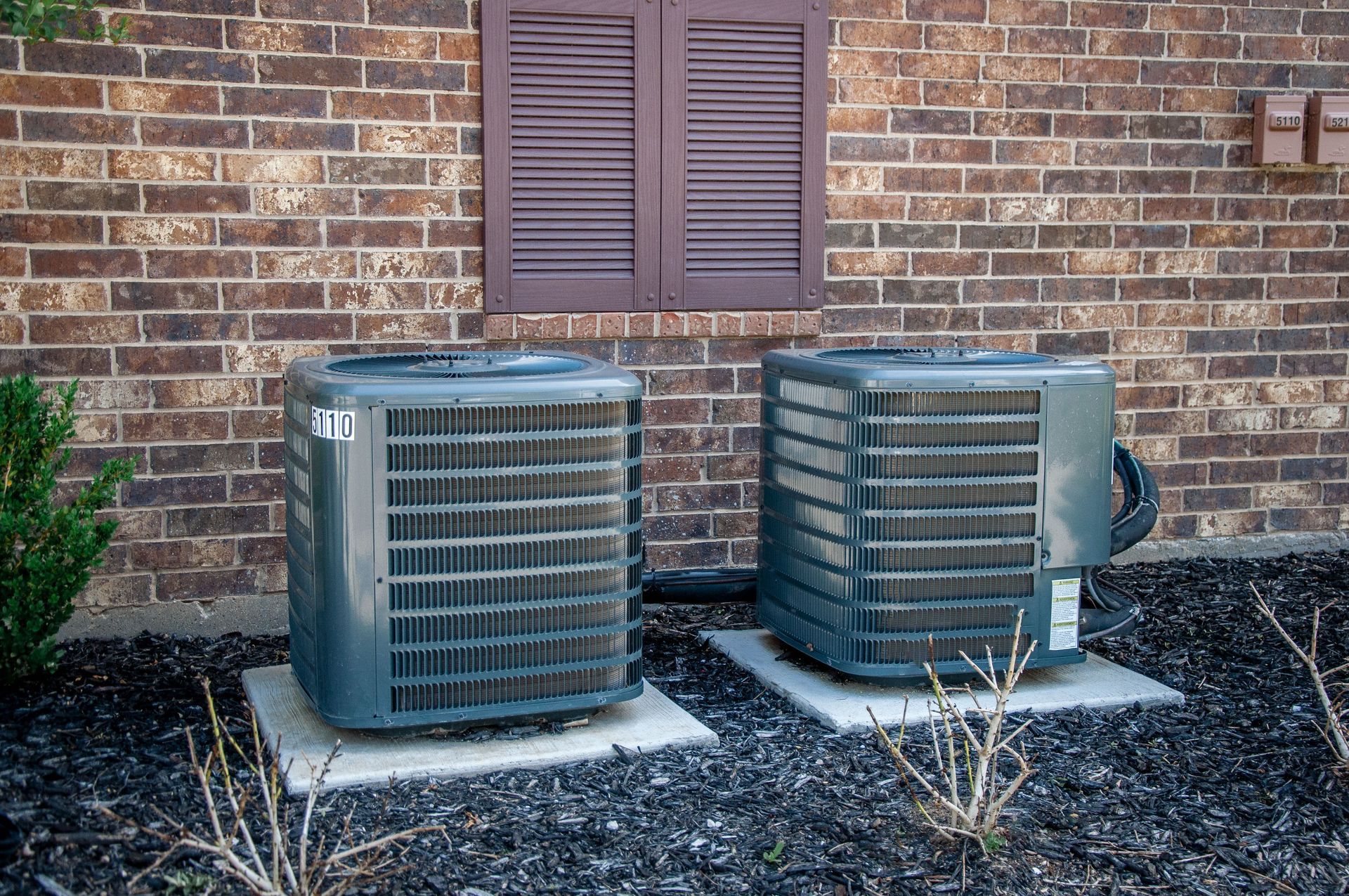Two outdoor air conditioner units beside a brick house wall and window shutters