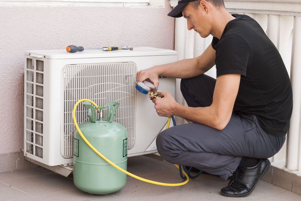 Technician kneeling beside outdoor AC unit, checking refrigerant with a hose and tank.