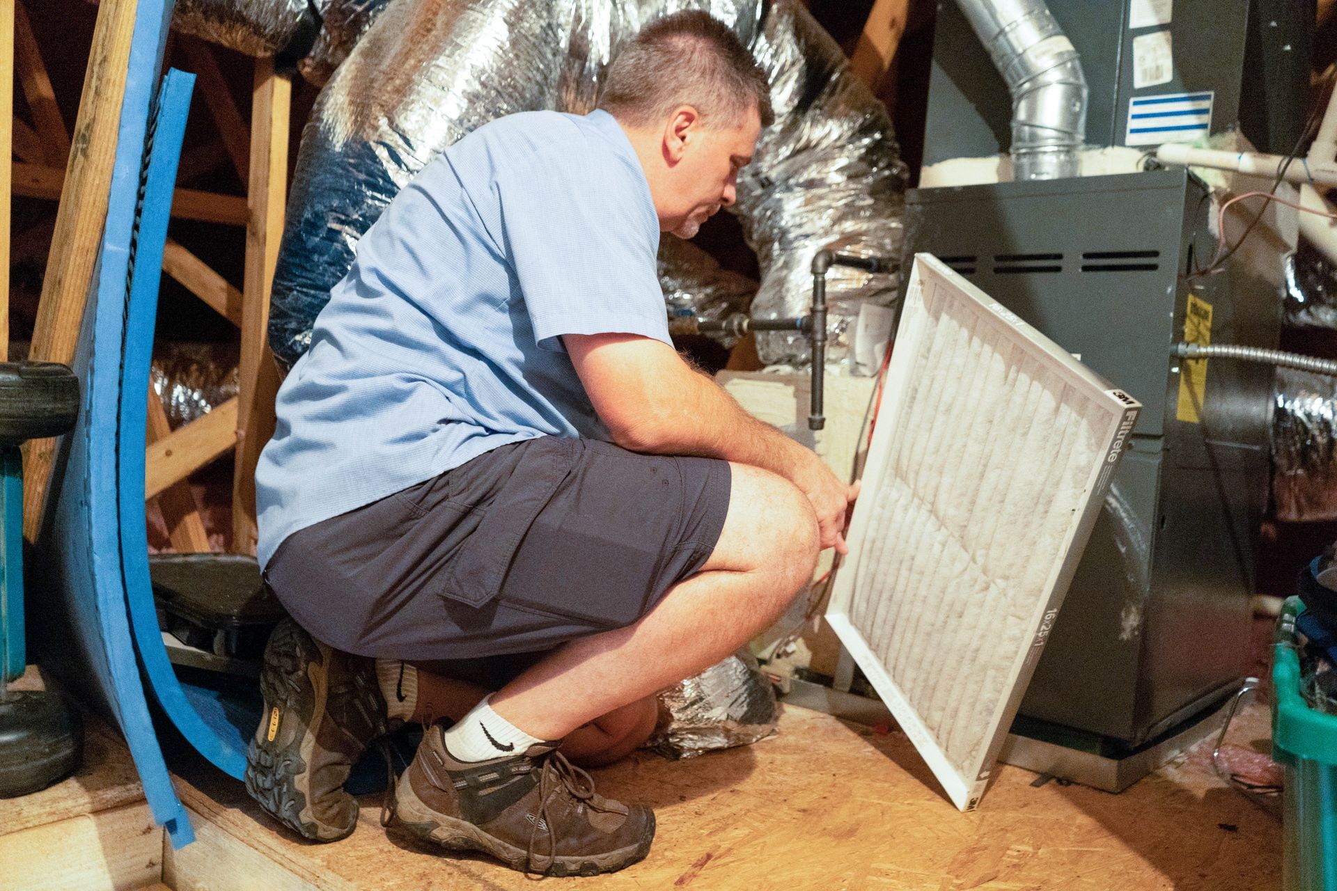 Technician crouching beside HVAC unit, checking a white air filter in a basement crawlspace