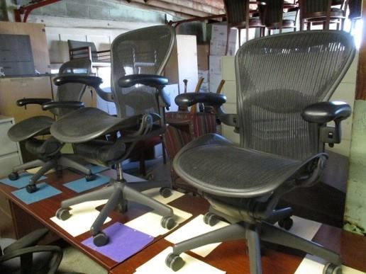 Four black mesh ergonomic office chairs on a wooden table, in a cluttered room.