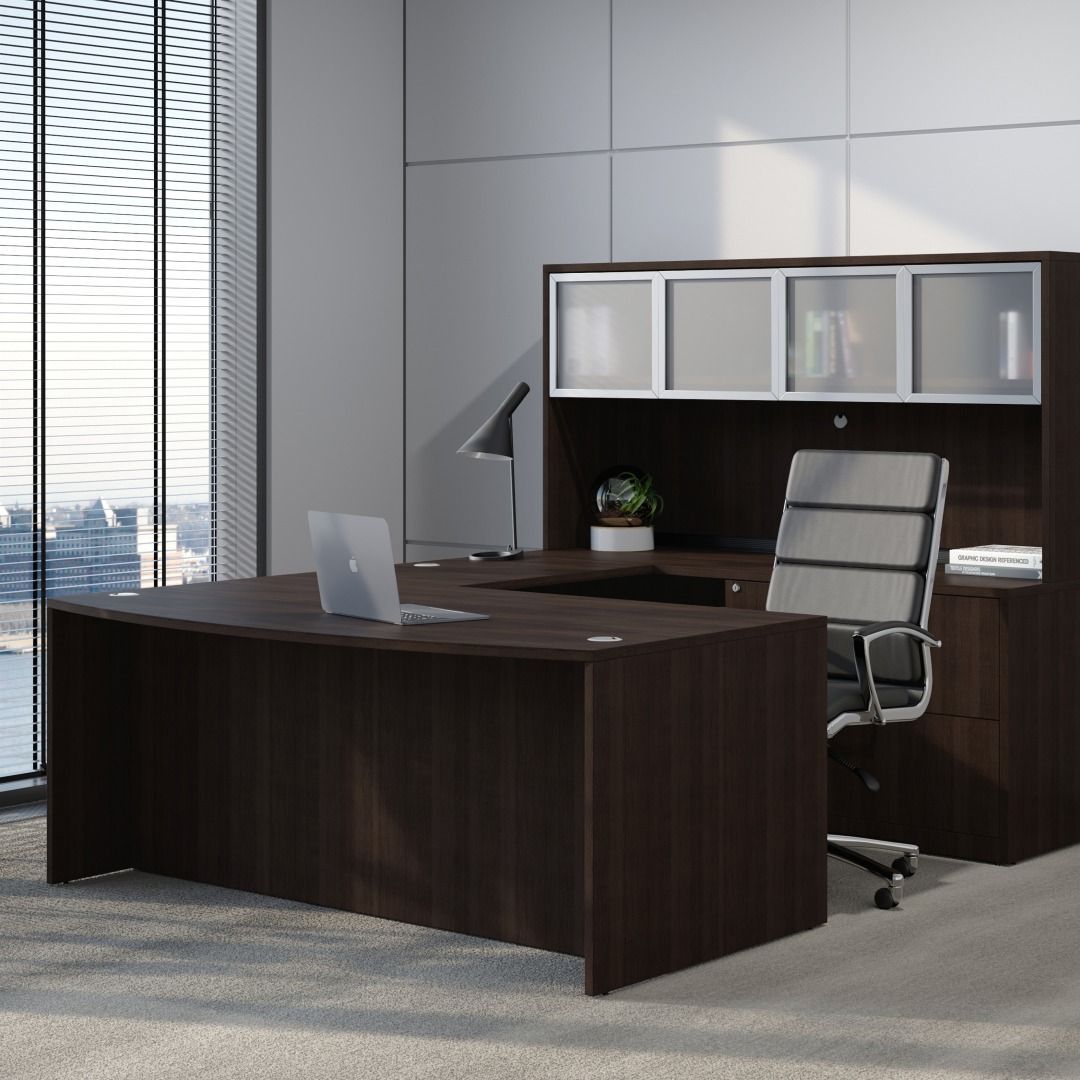 Dark brown office desk with computer, chair, and overhead cabinet. Window with cityscape.