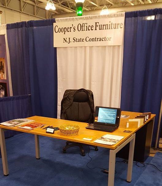 Office furniture booth with desk, laptop, chair, and sign. Blue curtains and a white backdrop are behind the desk.