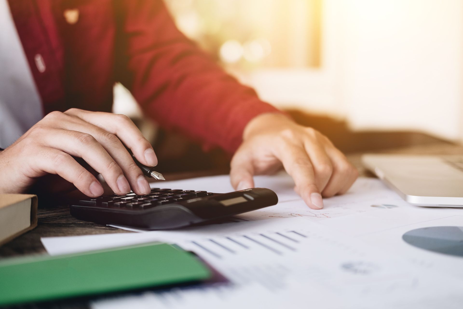 Person's hands using a calculator, looking at papers with graphs and a laptop on a desk.