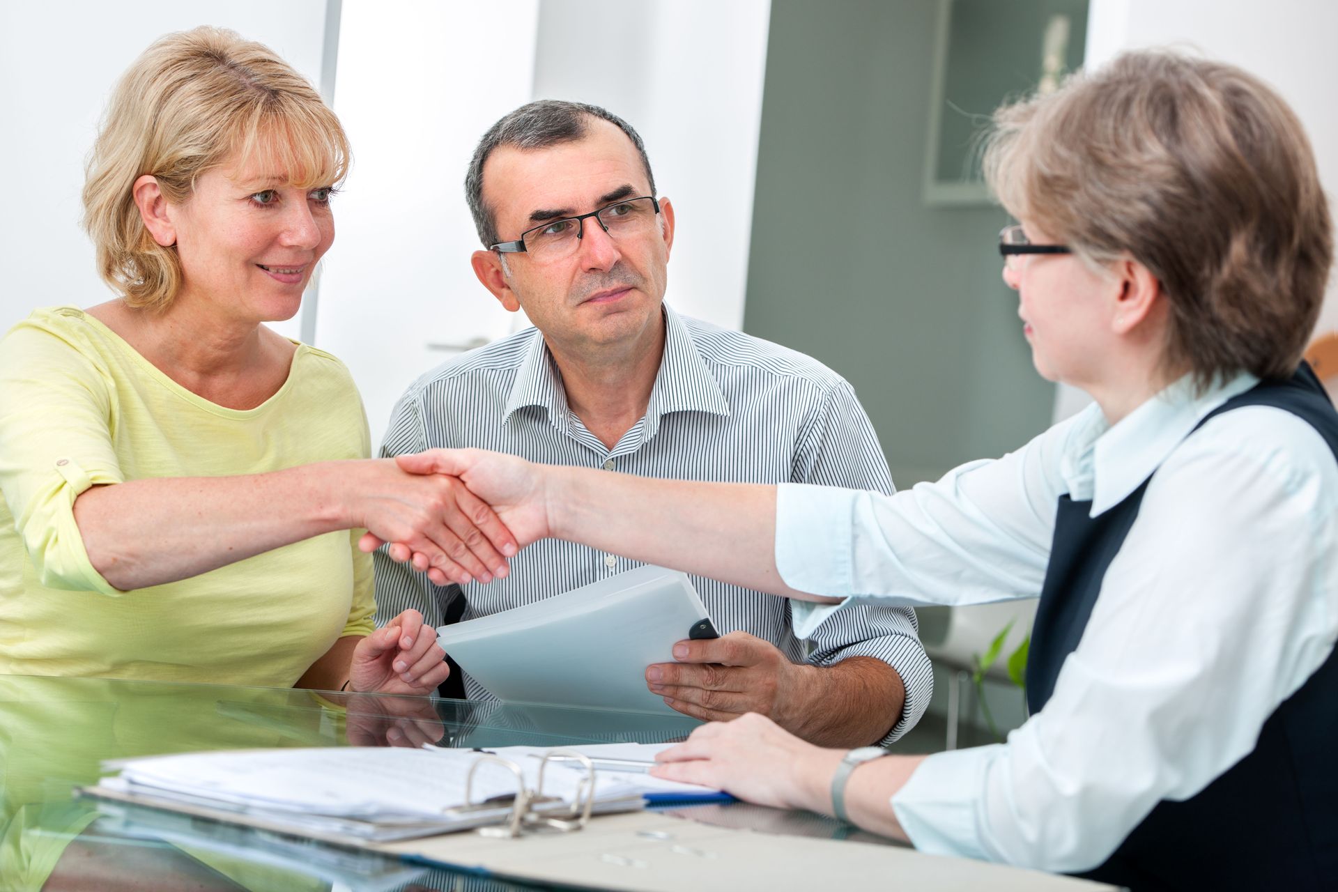 Woman shaking hands with another woman at a table; man watches. Indoors with paperwork.