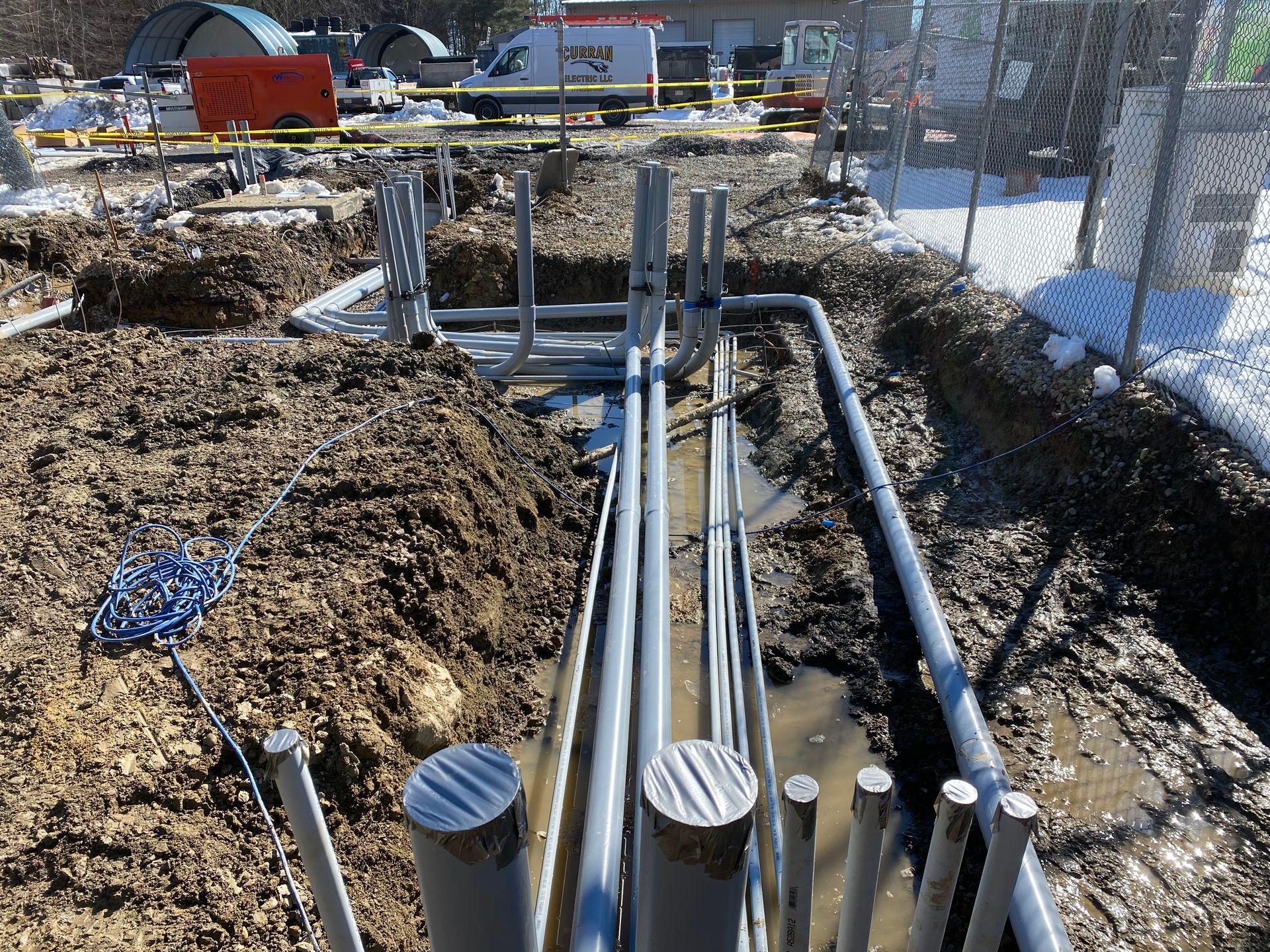 Trench dug in muddy ground, with gray pipes and metal supports, snow in the background.