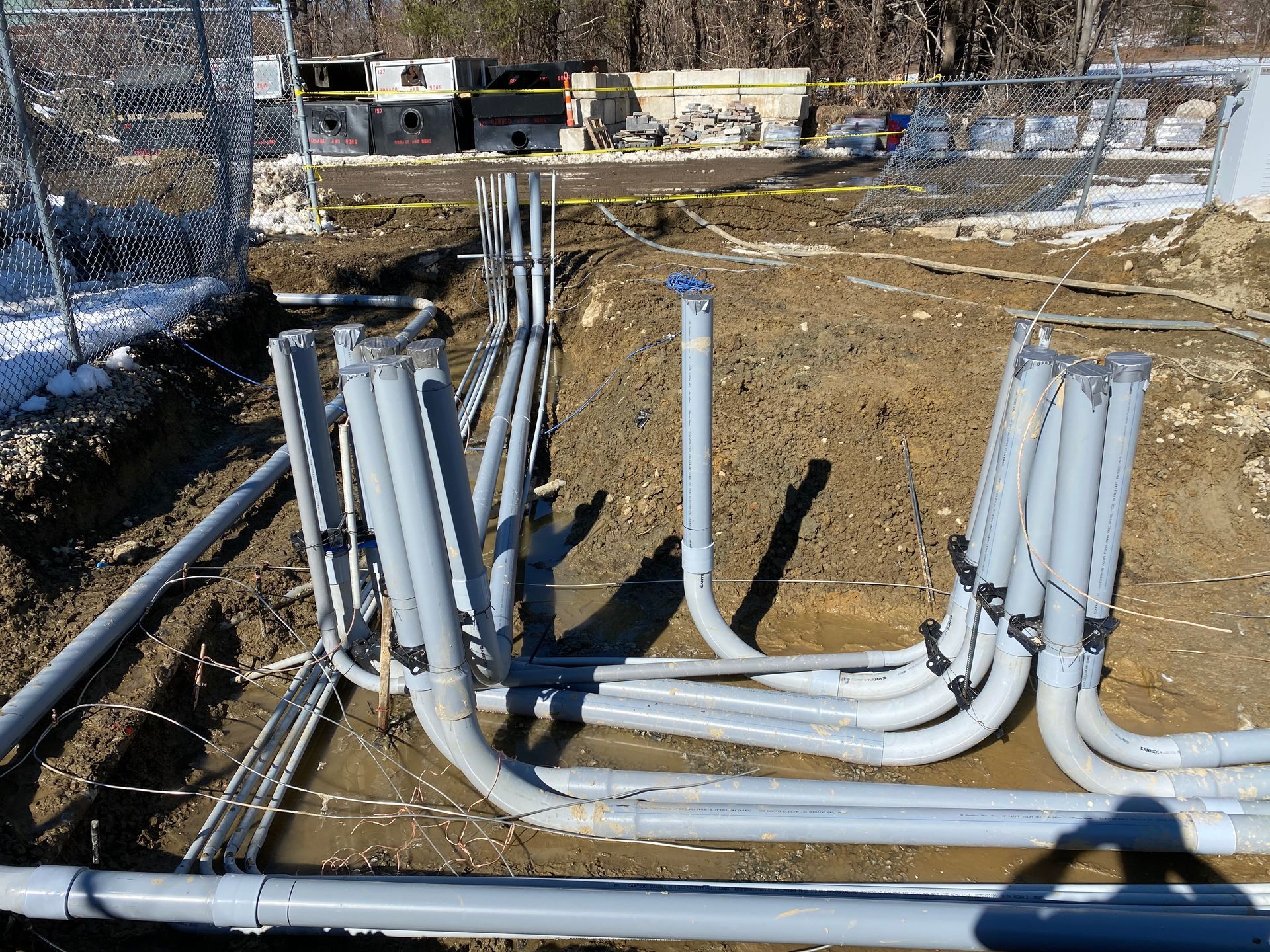 Gray electrical conduits in a trench, next to a wire fence. Construction site.