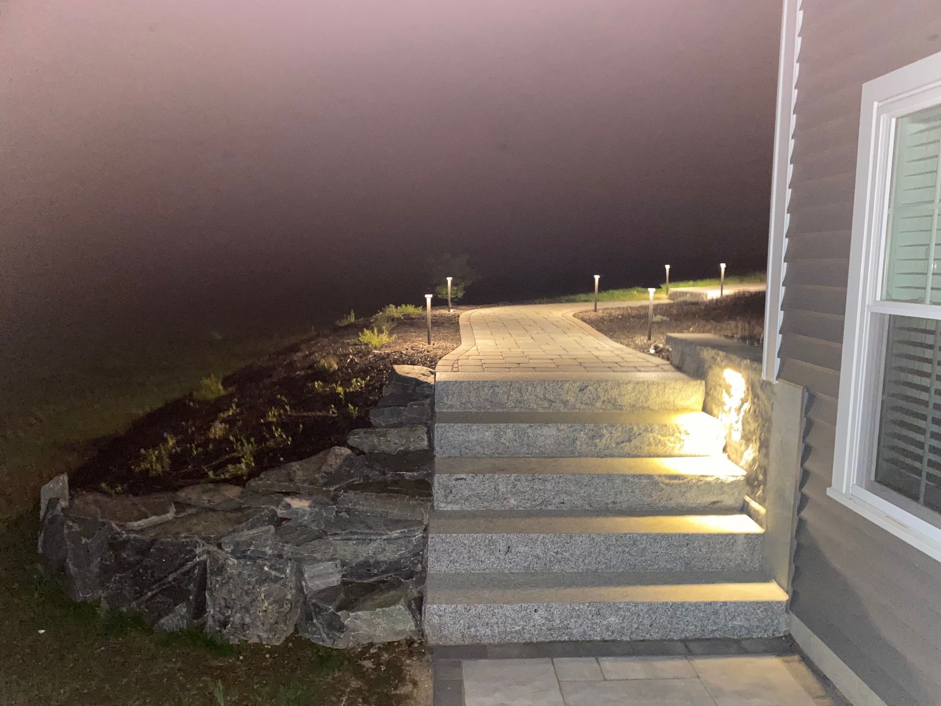 Concrete steps and pathway illuminated by lights lead uphill on a misty night.
