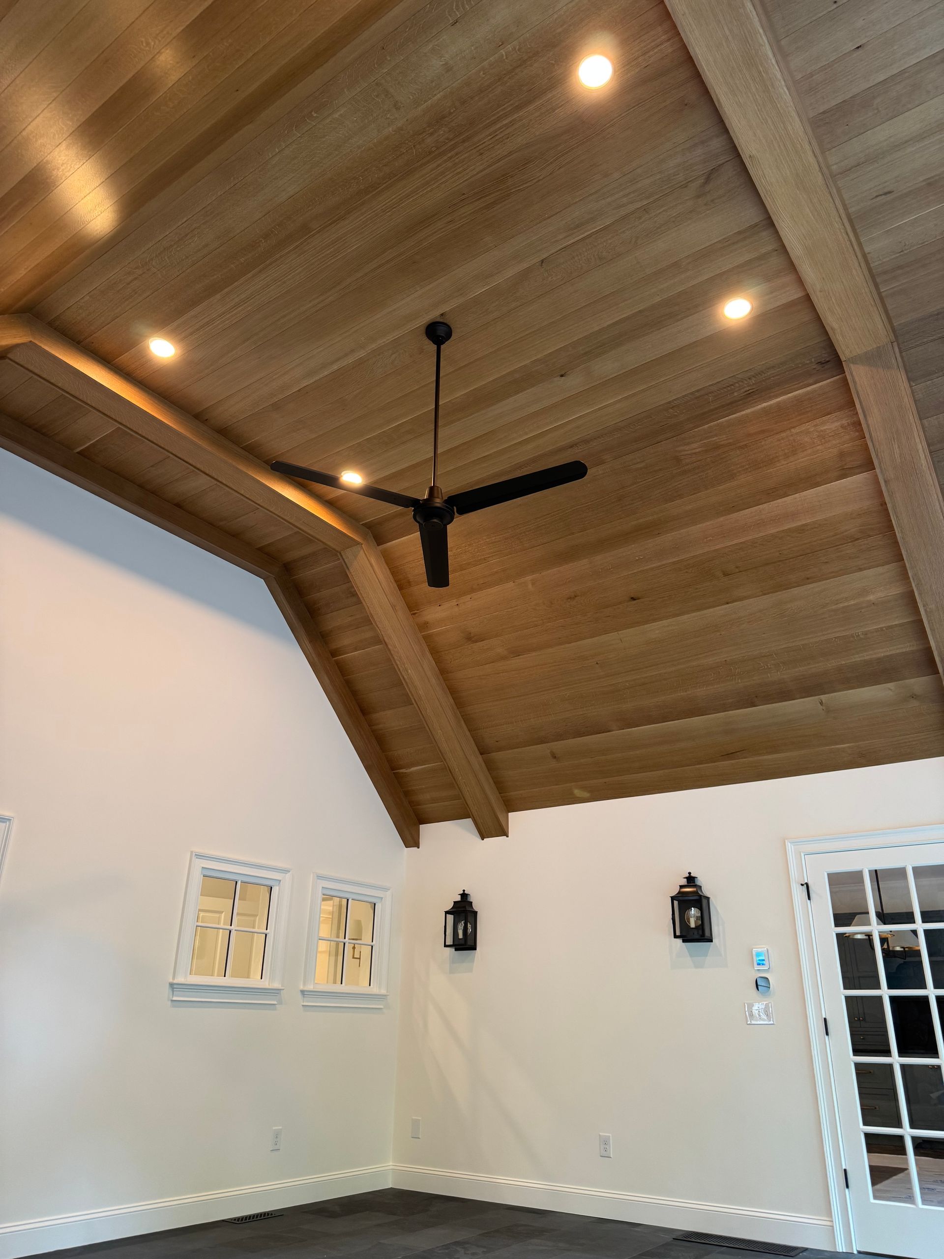 Wooden vaulted ceiling with recessed lights, black ceiling fan. Off-white walls, black light fixtures, windows.