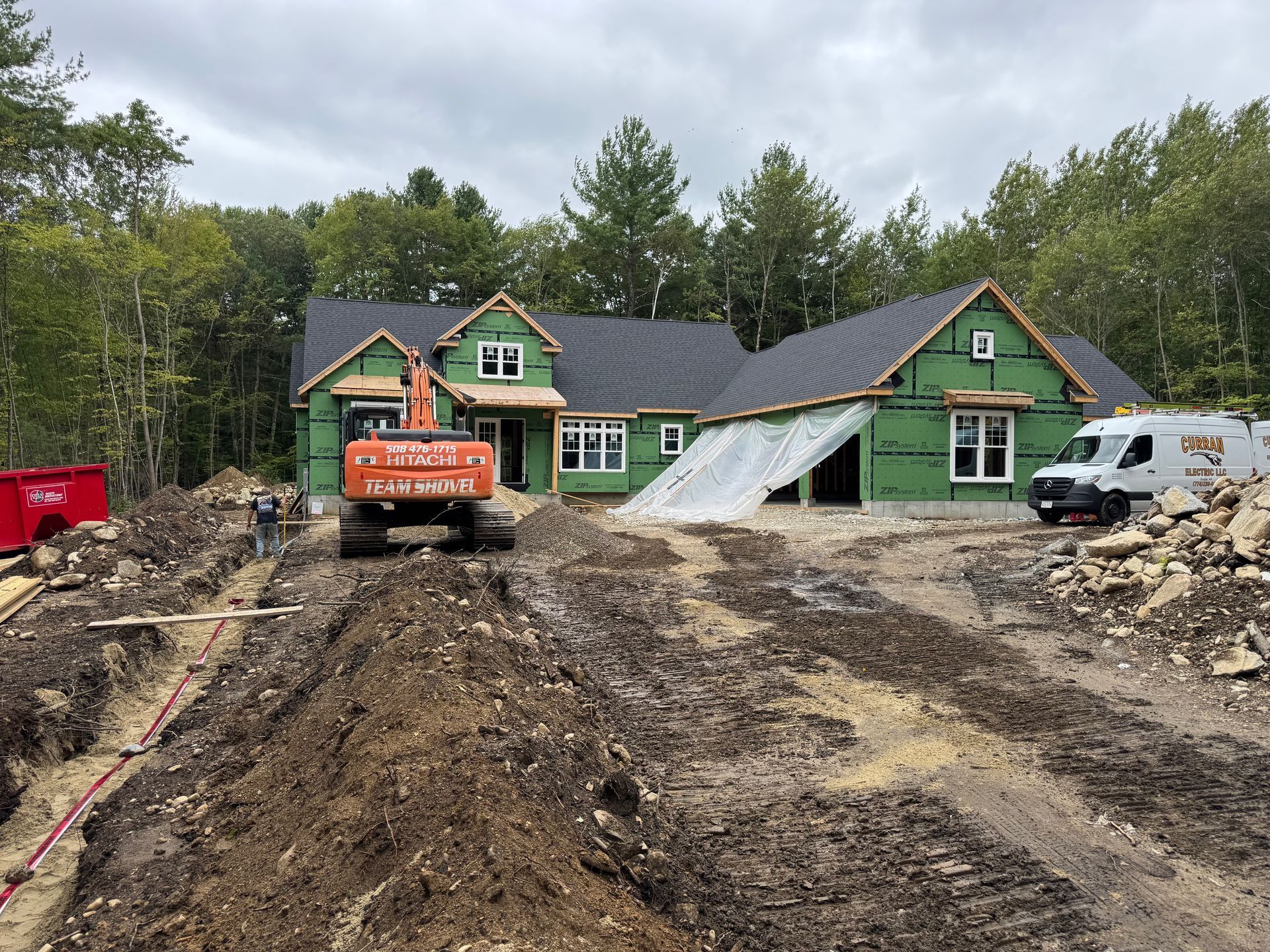 House under construction; excavator in front. Green siding, dark roof, trees in background, cloudy day.