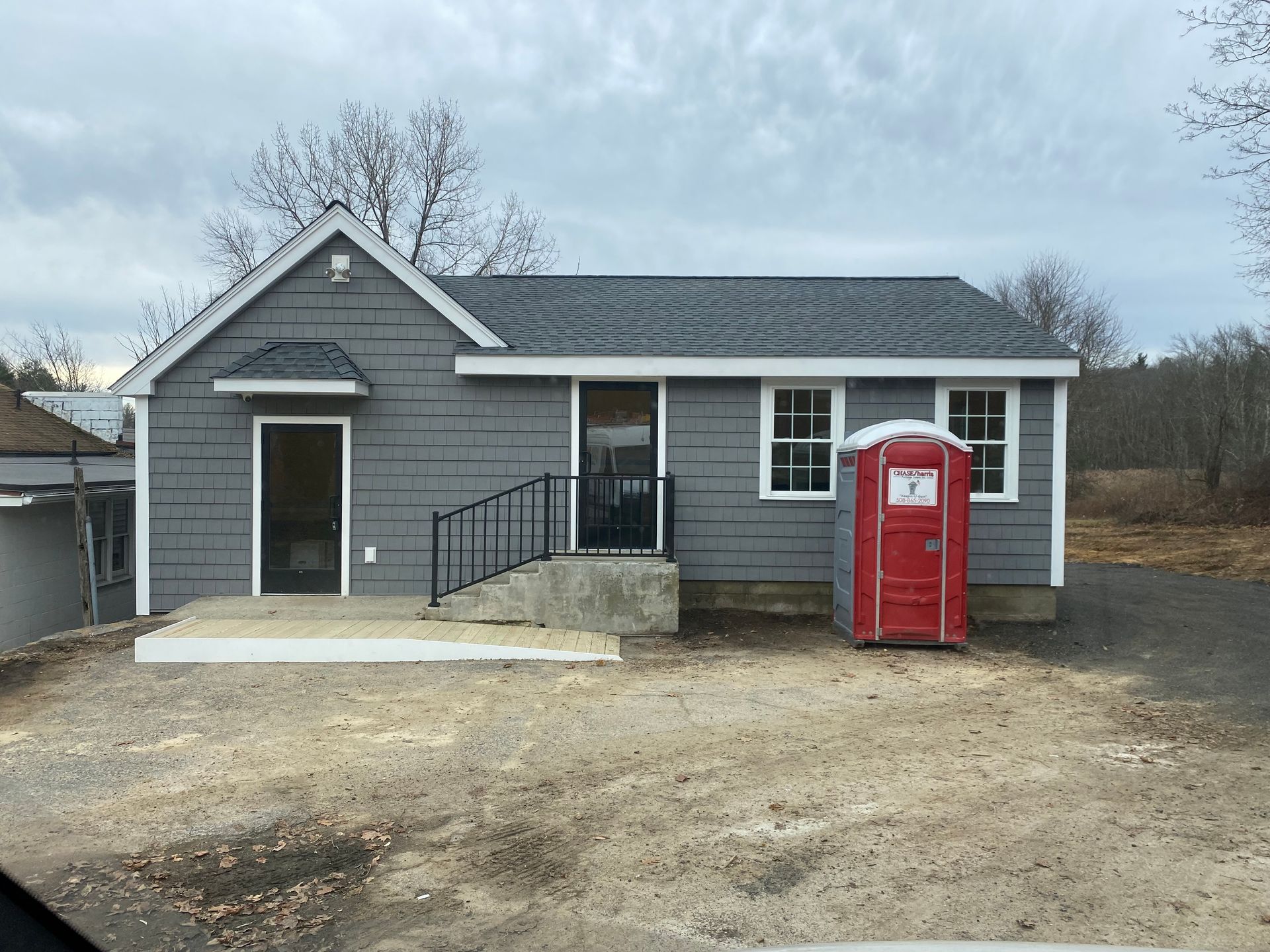 Small gray house with black railing, red portable toilet in front. Overcast day.