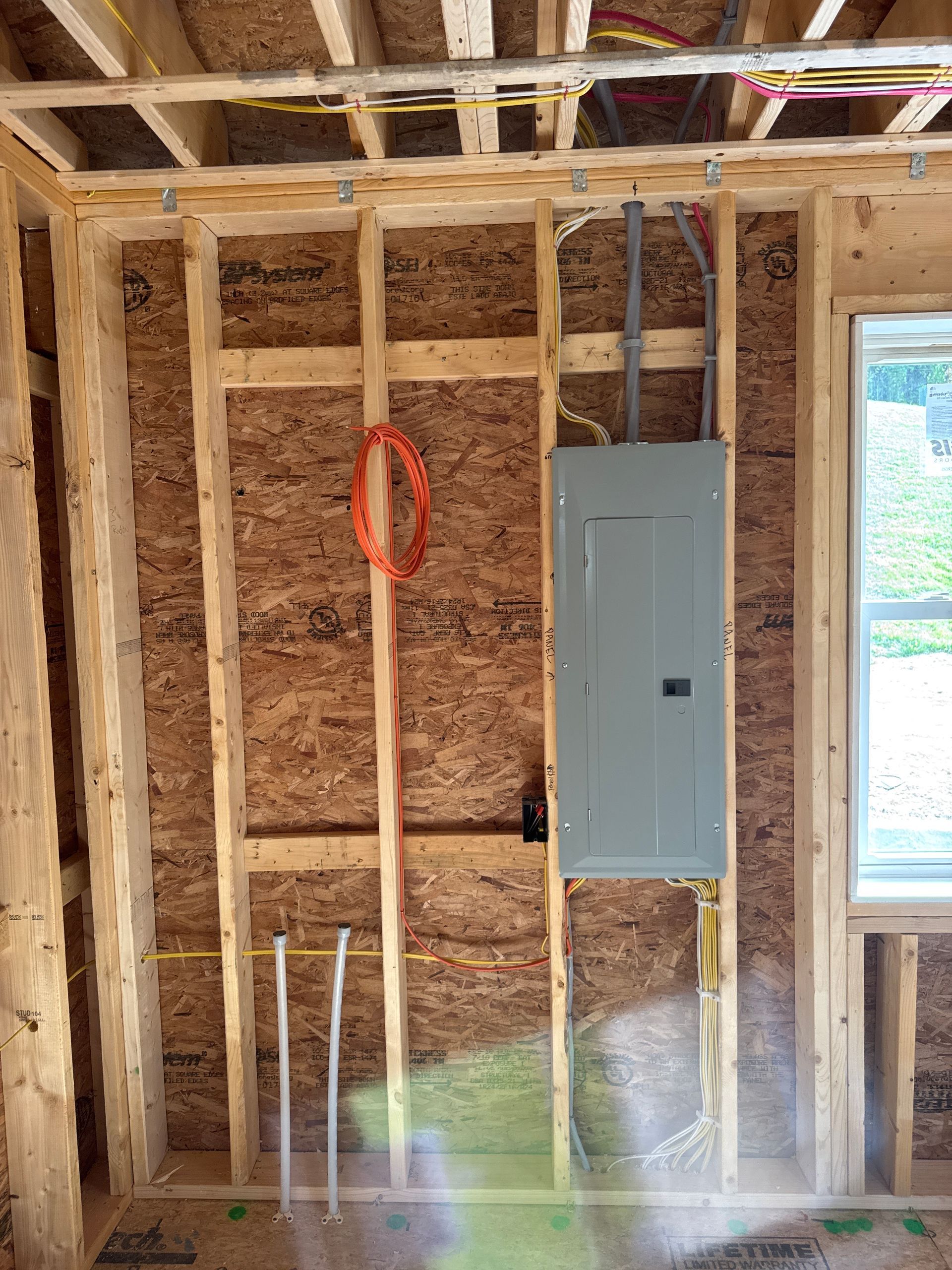 Electrical panel installed in a wooden wall frame during construction. Orange conduit and wiring visible.