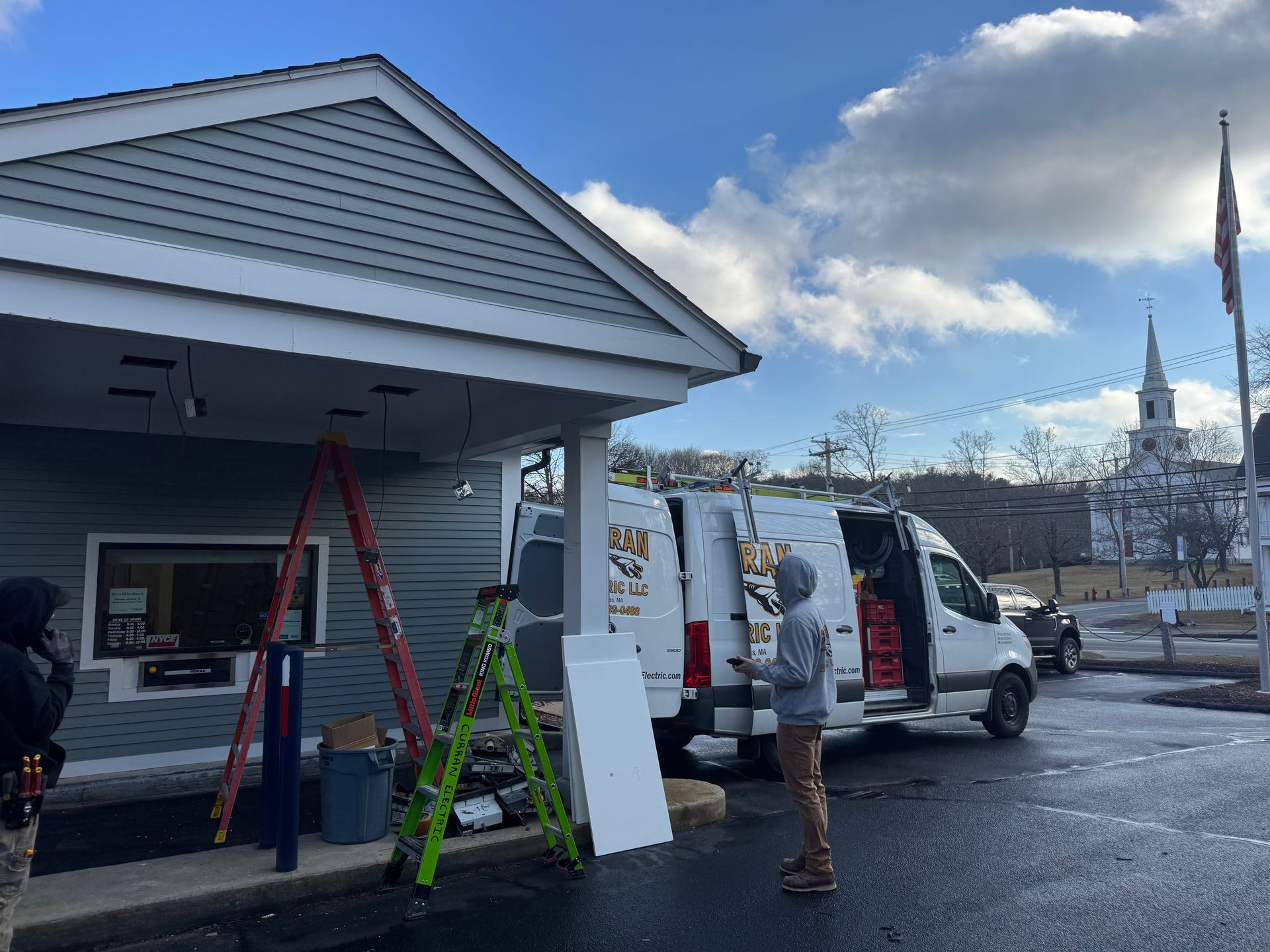 Workers installing exterior lights at a light blue building, near a white van and a church under a cloudy sky.