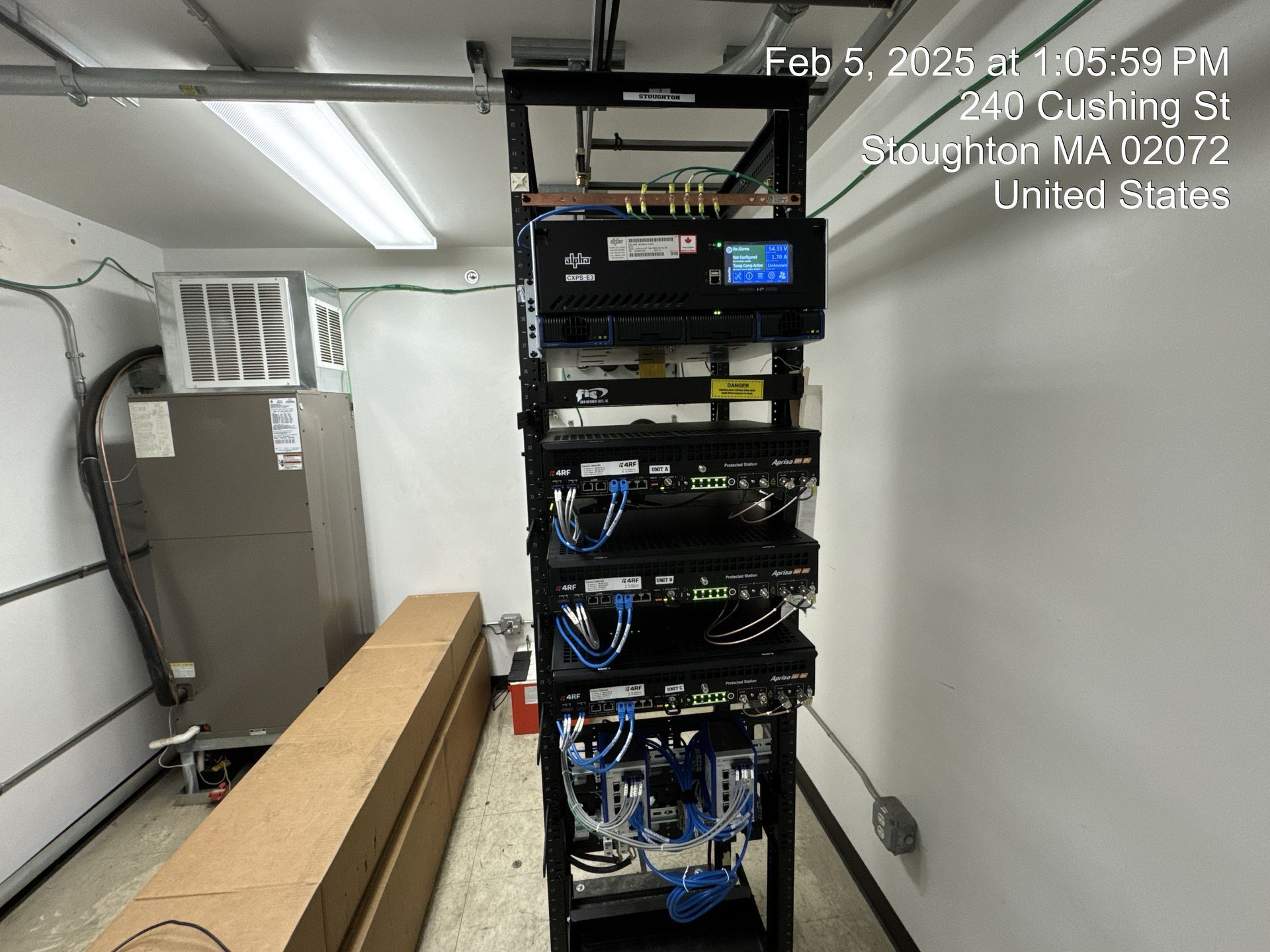 Equipment rack with several black electronic devices, wires, and a screen in a utility room.