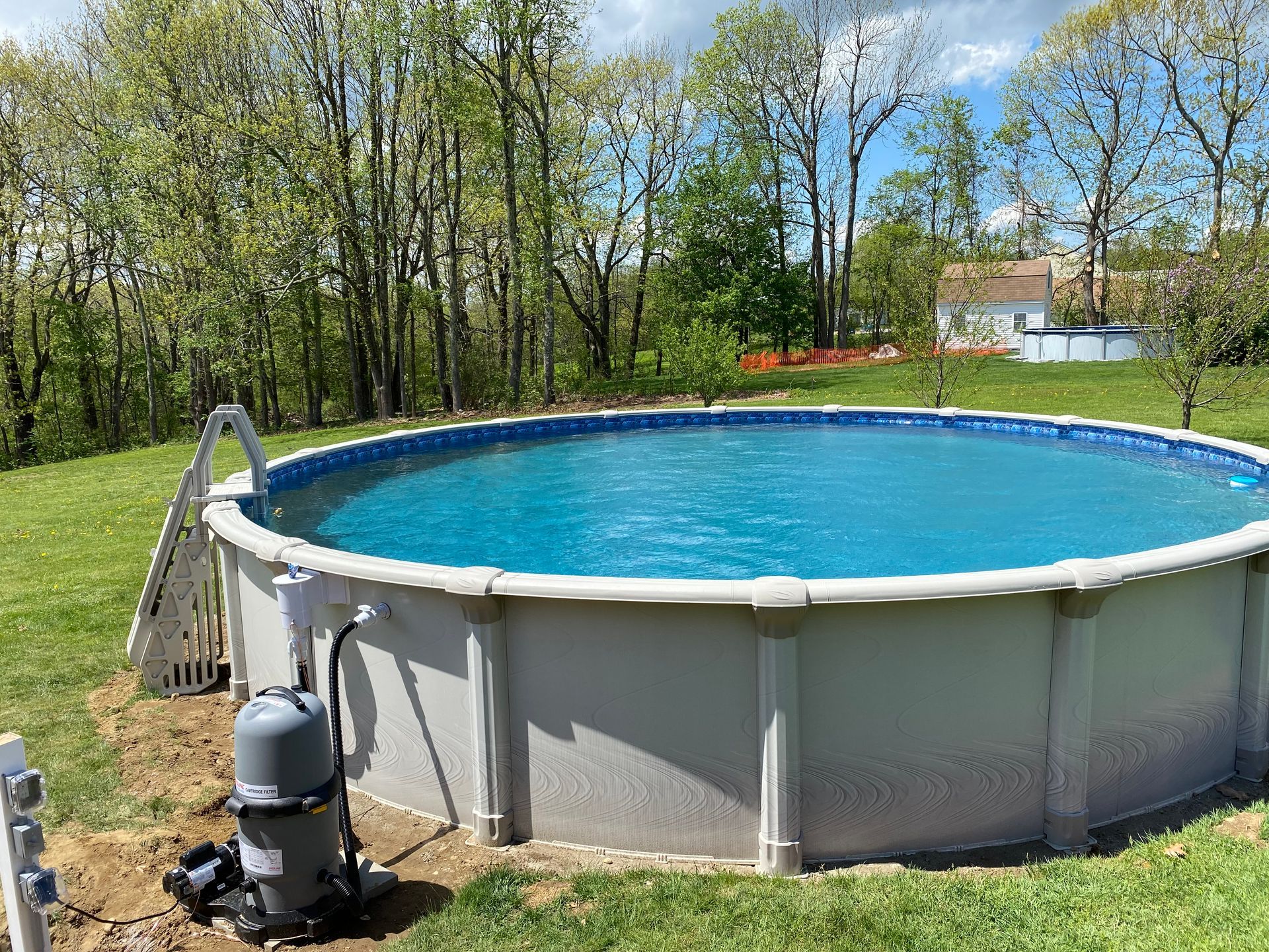 Above-ground swimming pool filled with blue water, surrounded by grass and trees. Filter system visible.