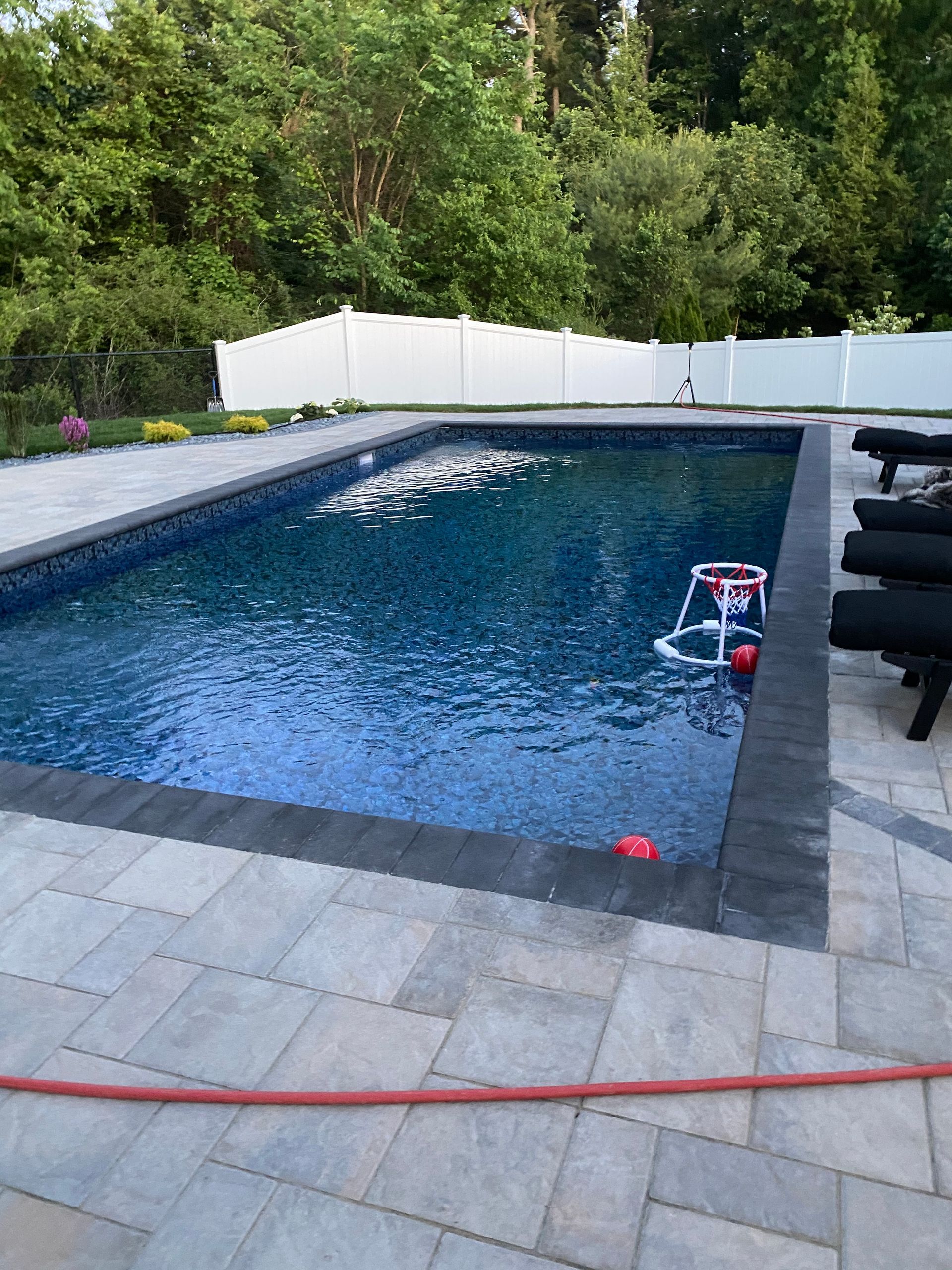 Rectangular pool with dark water, surrounded by pavers. A white fence and trees are in the background. A pool net game floats.