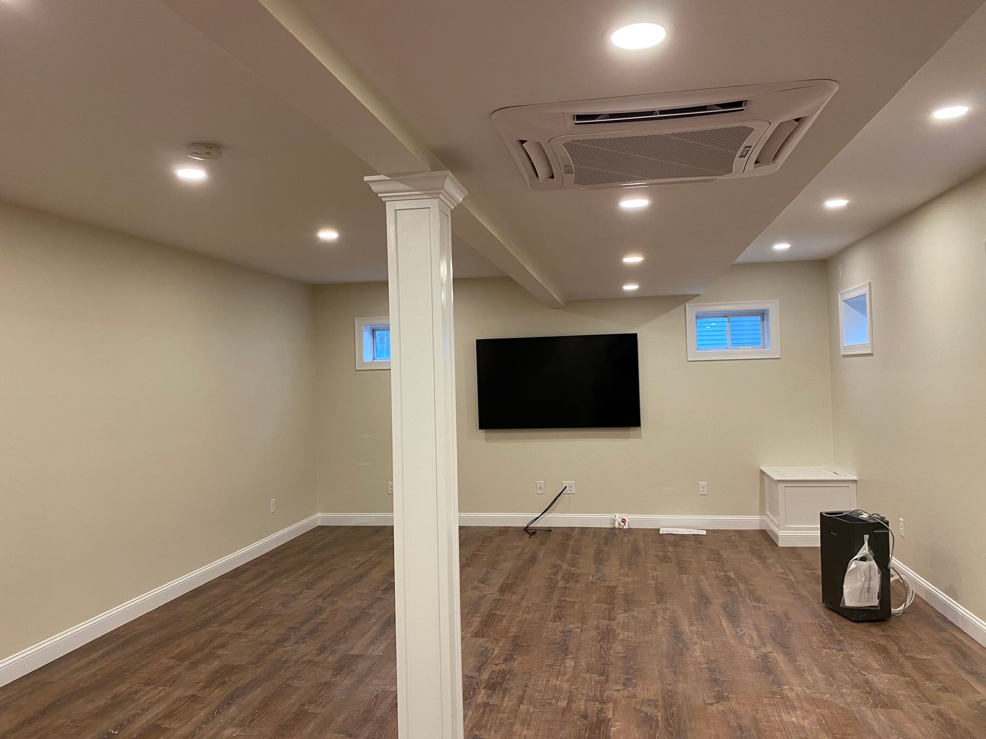 Finished basement with TV, ceiling lights, pillar, wood-look floor, and windows.